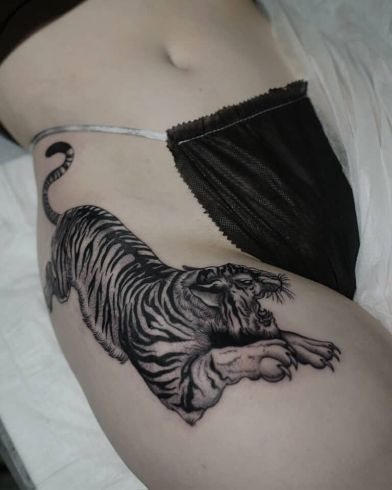 7sinstattoo.pl Osoba leży z misternym, czarno-szarym tatuażem warczącego tygrysa na biodrze i udzie. Ma na sobie czarną bieliznę i częściowo widoczny szary strój, prezentujący misterną pracę wykonaną przez studio tatuaży Poznań, uchwyconą na zdjęciu produktu DSC09992. Najlepsze studio tatuażu Seven Sins Tattoo