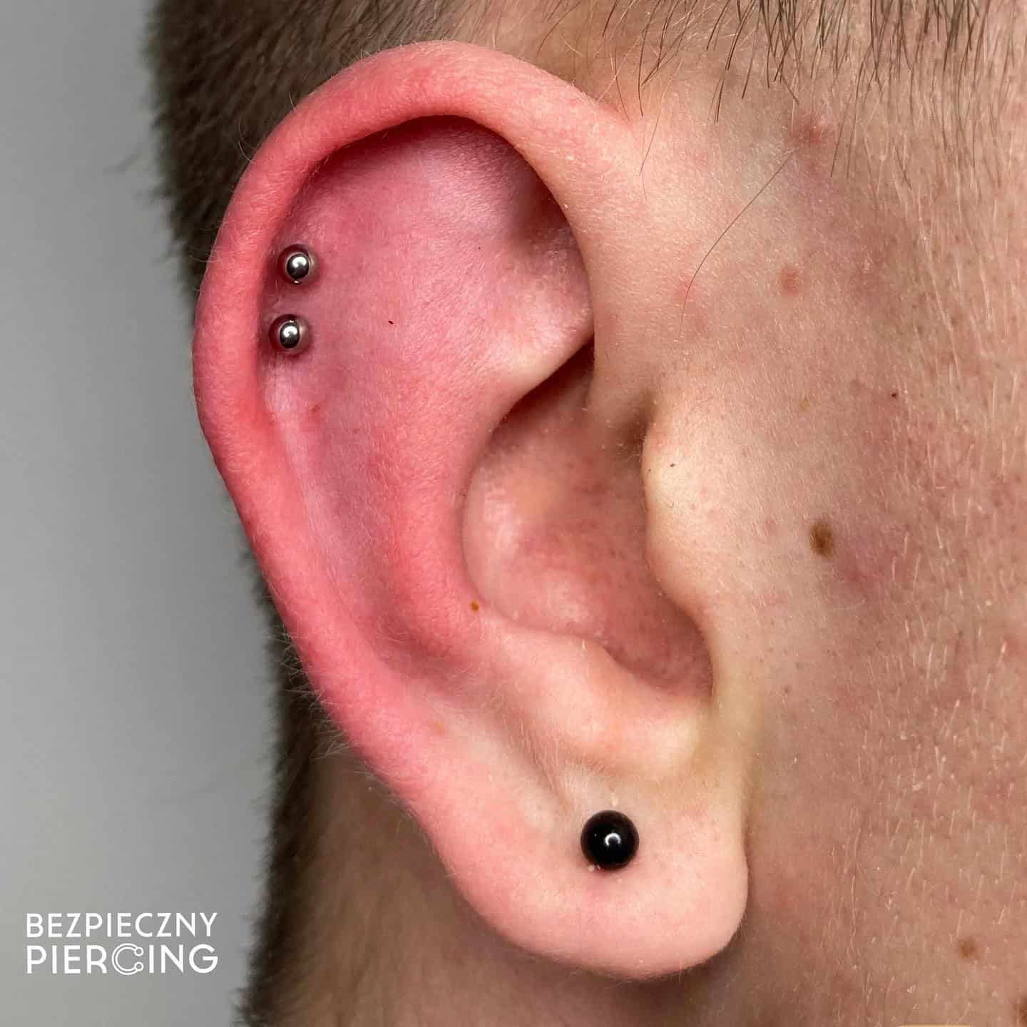 7sinstattoo.pl Zbliżenie ucha z dwoma piercingami: jeden w górnej części ucha z dwoma metalowymi kolczykami i drugi w płatku ucha z jednym czarnym kolczykiem. Skóra wokół ucha wydaje się gładka i zdrowa. „E79536A4-026C-44F3-BBFA-6E295AA29393” autorstwa najlepsze studio tatuażu Poznań znajduje się w lewym dolnym rogu. Najlepsze studio tatuażu Seven Sins Tattoo