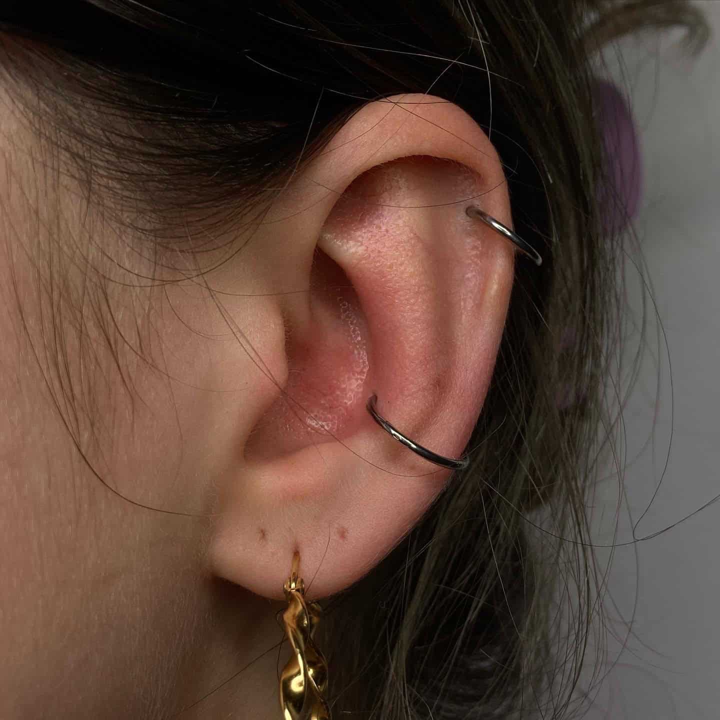 7sinstattoo.pl Zbliżenie ucha osoby z podwójnym przekłuciem helisy ozdobionym dwoma prostymi srebrnymi kółkami, uzupełnione przekłuciem płatka ucha ze złotym skręconym kolczykiem kółkiem. Włosy są schludnie schowane za uchem, odsłaniając jasną skórę. Odkryj podobne style i bardziej kreatywne przekłucia w Studio Tatuażu Poznań, gdzie znajdziesz produkty takie jak F1AF7148-6E9F-4C2F-98B5-419D6172282E. Najlepsze studio tatuażu Seven Sins Tattoo