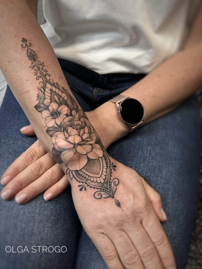 7sinstattoo.pl Przedramię prezentujące IMG-5630, misterny czarno-szary kwiatowy tatuaż mandali rozciągający się na dłoń i nadgarstek. Osoba jest ubrana w biały top, niebieskie dżinsy i srebrny smartwatch. Ten misterny tatuaż, wykonany w Najlepszym Studio Tatuażu Poznań, zawiera szczegółowe kwiaty i ozdobne wzory z tekstem „OLGA STROGO. Najlepsze studio tatuażu Seven Sins Tattoo
