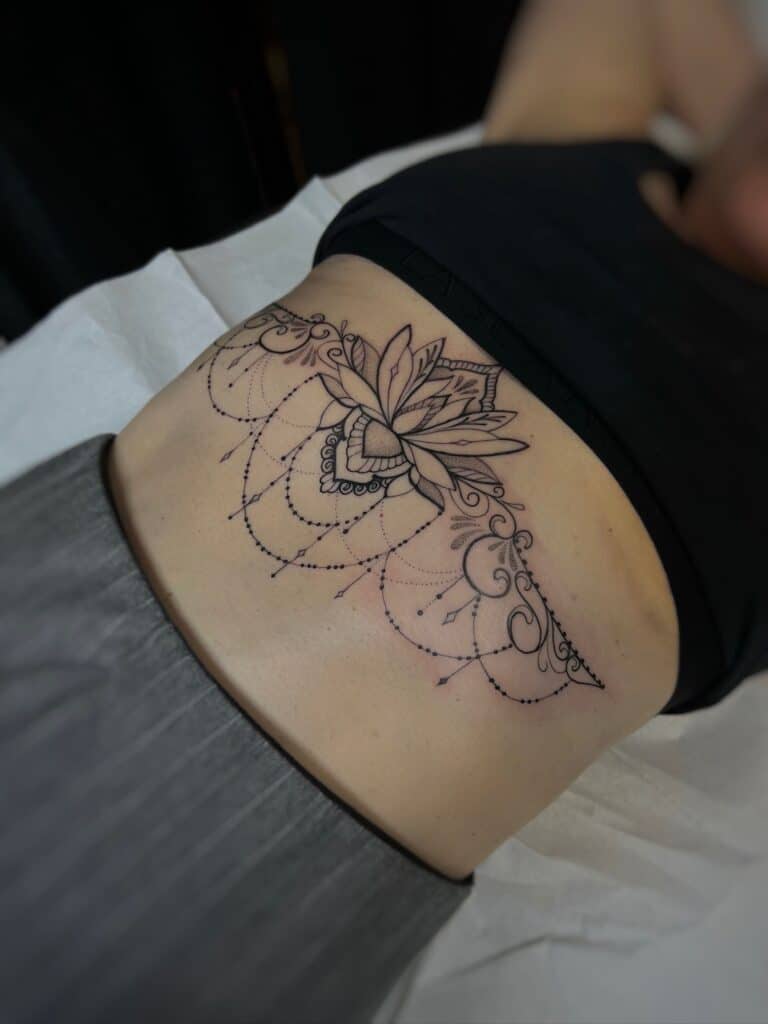 7sinstattoo.pl W IMG-5736 szczegółowe zbliżenie ukazuje dolną część pleców osoby ozdobioną misternym tatuażem z cenionego studia tatuażu Poznań. Tatuaż przedstawia widoczny kwiat lotosu w centrum, otoczony misternymi, symetrycznymi wzorami i precyzyjnymi kropkami, które rozciągają się na dolnej części pleców. Osoba jest umieszczona na białym prześcieradle. Najlepsze studio tatuażu Seven Sins Tattoo