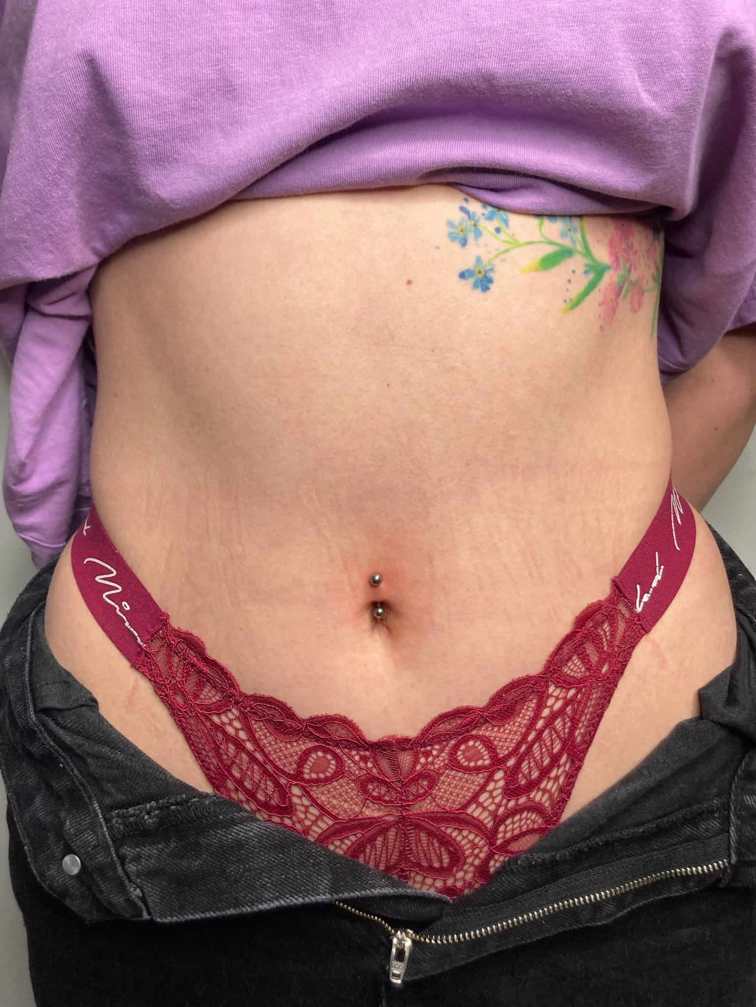 7sinstattoo.pl Zbliżenie na dolną część brzucha i talię osoby ukazuje czerwone koronkowe stringi wystające z rozpiętych czarnych dżinsów. Osoba ta ma piercing w pępku i może pochwalić się żywym kwiatowym tatuażem ze Studio Tatuażu Poznań na boku. Ma na sobie lekko uniesioną fioletową koszulę, uchwyconą na zdjęciu o nazwie IMG_0189. Najlepsze studio tatuażu Seven Sins Tattoo