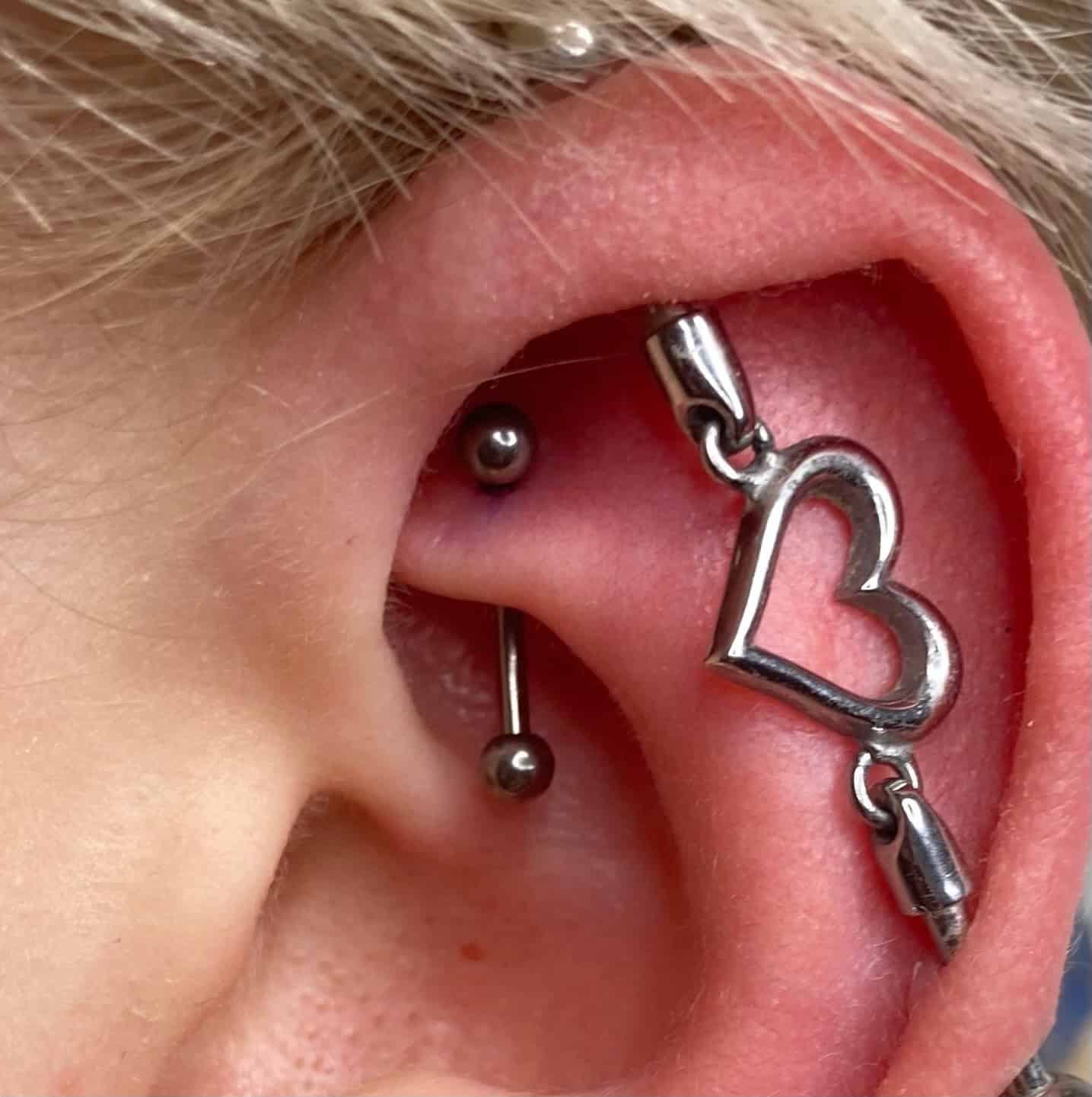 7sinstattoo.pl Zbliżenie ukazuje ucho osoby ozdobione wieloma piercingami, w tym industrialnym piercingiem z metalowym prętem. Dodatkowo, zwisający kolczyk z zawieszką w kształcie serca zwisa z górnej części ucha. Blond włosy osoby podkreślają odważny styl, idealny do zaprezentowania w najlepszym studio tatuażu w Poznaniu. Ta scena doskonale oddaje IMG_0646. Najlepsze studio tatuażu Seven Sins Tattoo