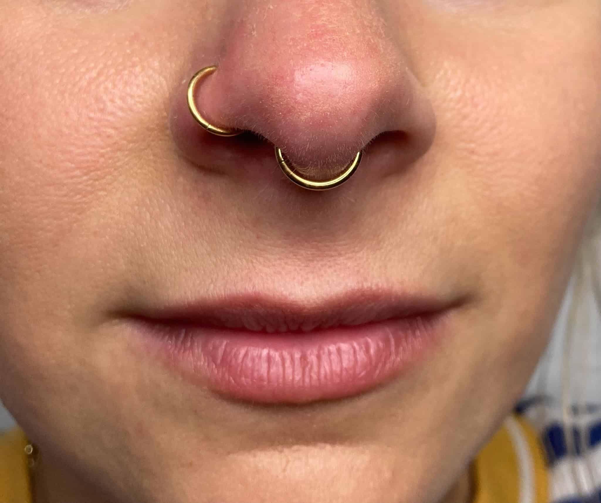 7sinstattoo.pl Zbliżenie dolnej części twarzy osoby z obrazu IMG_0658, ukazujące dwa złote kolczyki w nosie; jeden zdobiący lewe nozdrze, a drugi przewleczony przez przegrodę nosową. Osoba ma różowe usta i jasną skórę. Nieco rozmyte tło sugeruje otoczenie przypominające studio tatuaży Poznań, z nutami niebiesko-białego wzorzystego materiału. Najlepsze studio tatuażu Seven Sins Tattoo