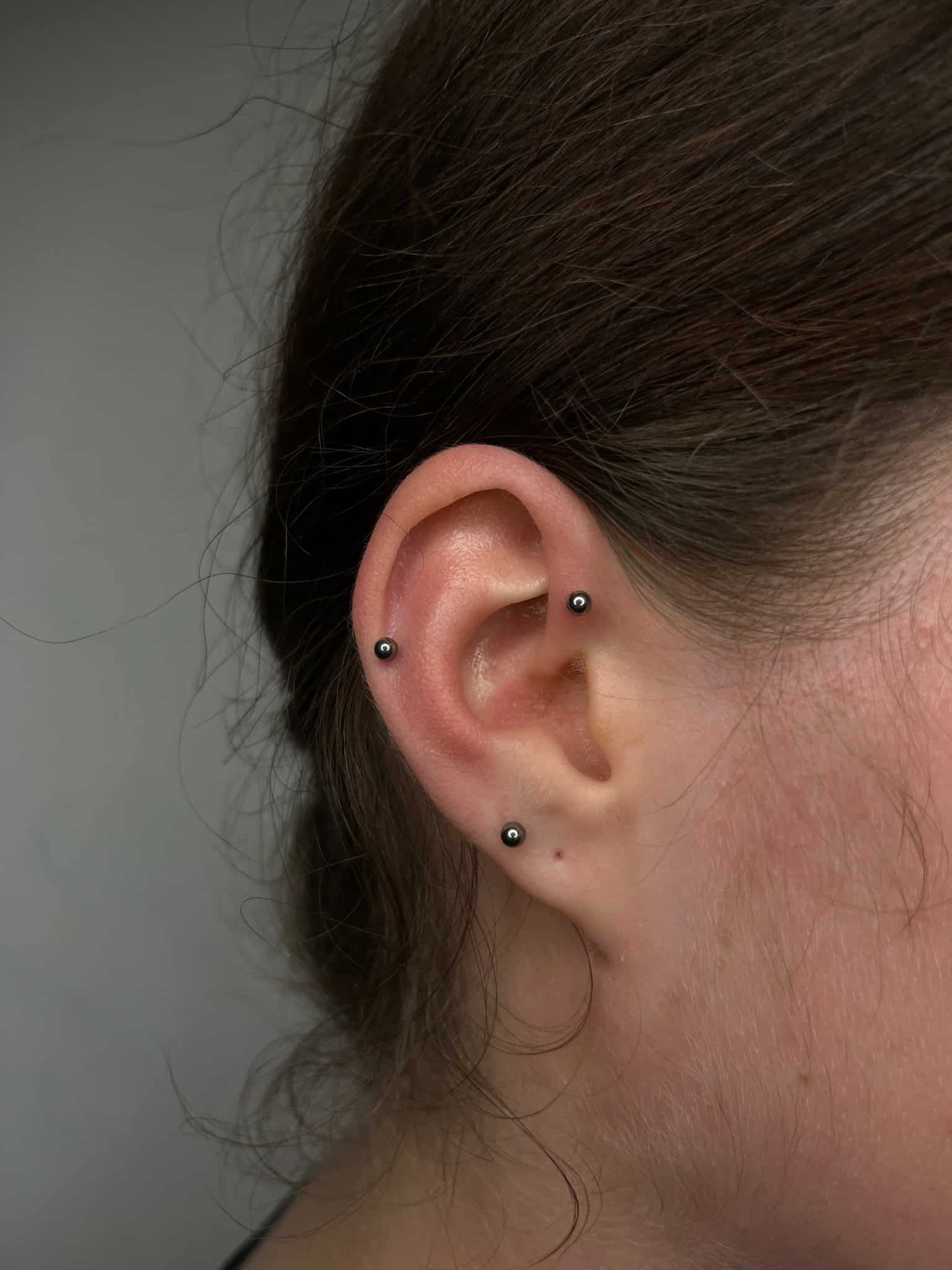 7sinstattoo.pl Zbliżenie ucha osoby z dwoma małymi metalowymi kolczykami w kształcie kulek: jednym na górnej chrząstce i jednym na płatku ucha. Włosy są związane z tyłu, wyraźnie odsłaniając ucho na neutralnym tle w IMG_0741, najlepsze studio tatuażu Poznań. Najlepsze studio tatuażu Seven Sins Tattoo