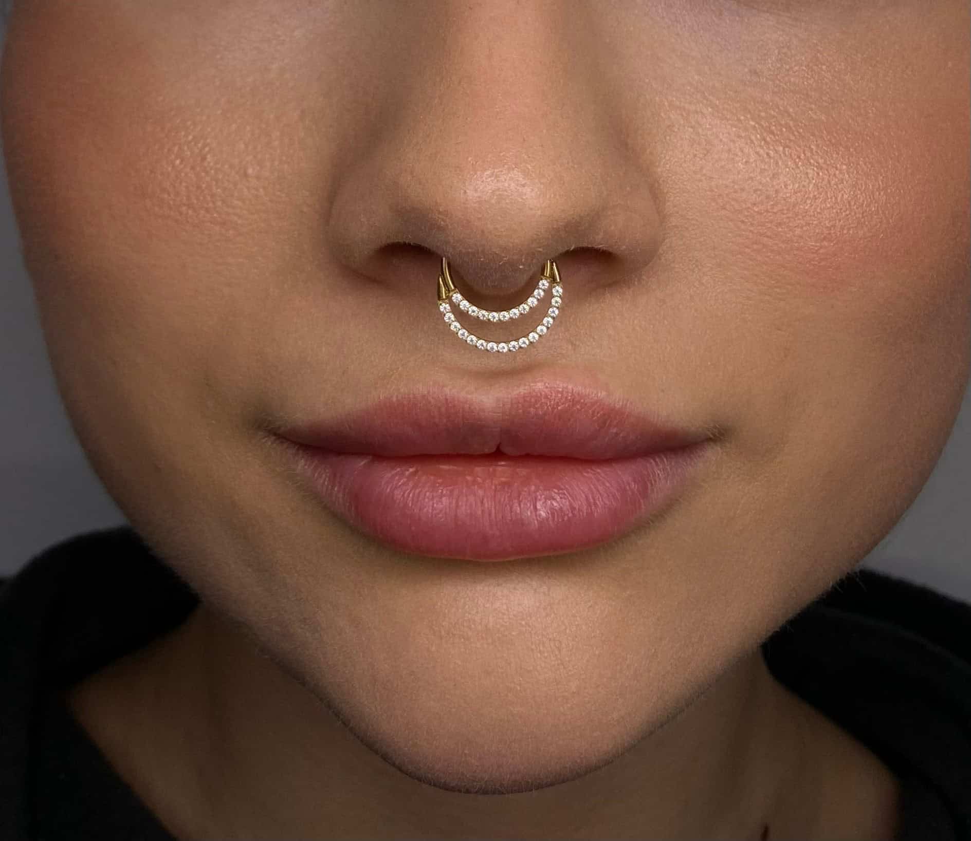 7sinstattoo.pl Zbliżenie twarzy osoby, skupiające się na nosie i ustach. Nosi ona podwójny pierścień przegrody nosowej IMG_0809 ozdobiony małymi, białymi kamieniami szlachetnymi z najlepszego studia tatuażu Poznań. Pierścień przegrody nosowej ma dwie równoległe linie kamieni szlachetnych w kształcie półksiężyca. Osoba ma gładką, lekko opaloną skórę i pełne usta. Najlepsze studio tatuażu Seven Sins Tattoo