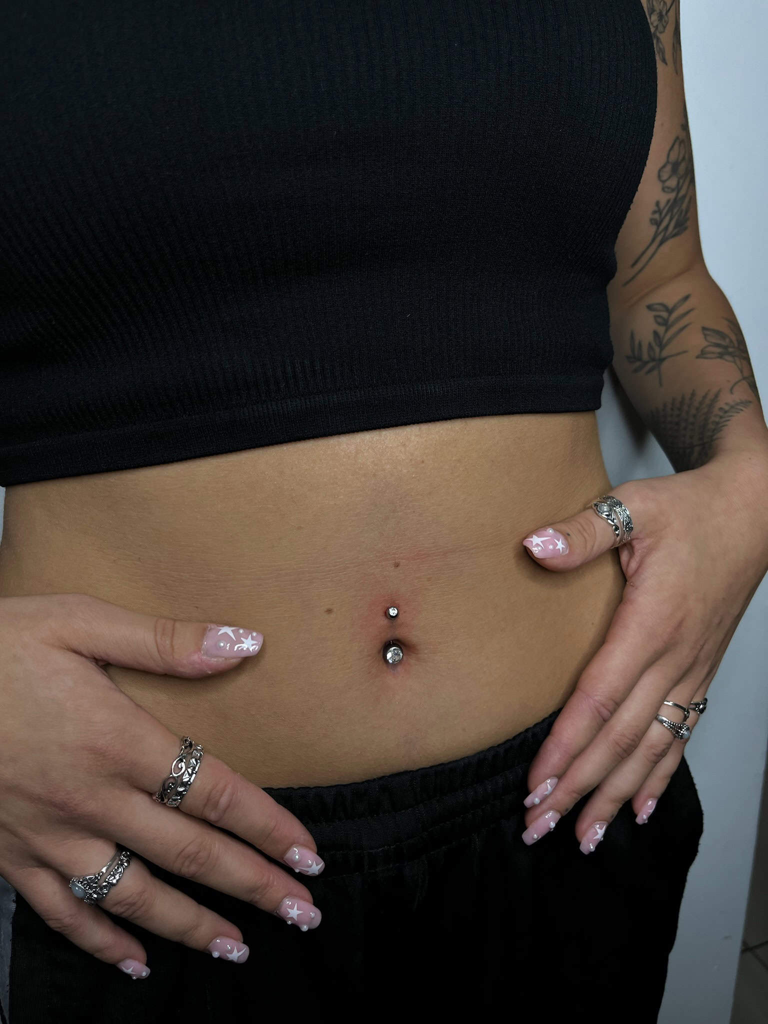 7sinstattoo.pl Osoba pewnie eksponuje piercing pępka ozdobiony dwoma srebrnymi ćwiekami, mając na sobie czarny krótki top i czarne spodnie. Dłonie tej osoby, ozdobione pierścionkami i zadbane paznokcie z jasnoróżowym lakierem i kwiatowymi wzorami, odsłaniają widoczne tatuaże na przedramieniu ze studia tatuażu Poznań. Ten uderzający obraz został uchwycony w produkcie IMG_0836. Najlepsze studio tatuażu Seven Sins Tattoo