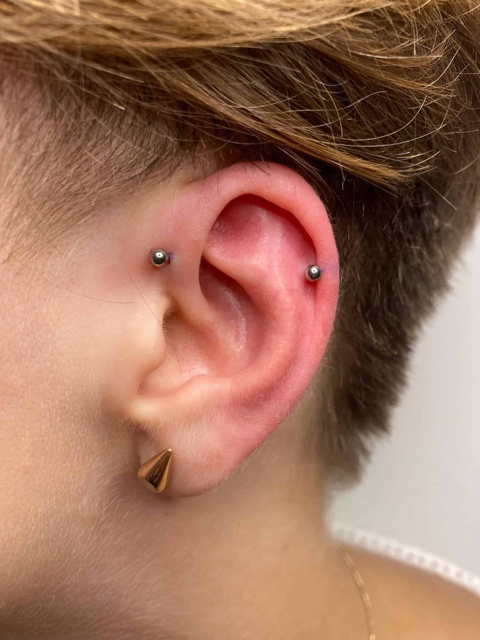 7sinstattoo.pl Zbliżenie ucha osoby z dwoma przekłuciami wewnętrznej chrząstki i przekłuciem helix, wszystkie z małymi, srebrnymi metalowymi ćwiekami (IMG_0837). Osoba ma również złoty, trójkątny kolczyk w płatku ucha. Z krótkimi, jasnobrązowymi włosami prezentuje swój styl inspirowany klimatem studia tatuaży Poznań. Najlepsze studio tatuażu Seven Sins Tattoo
