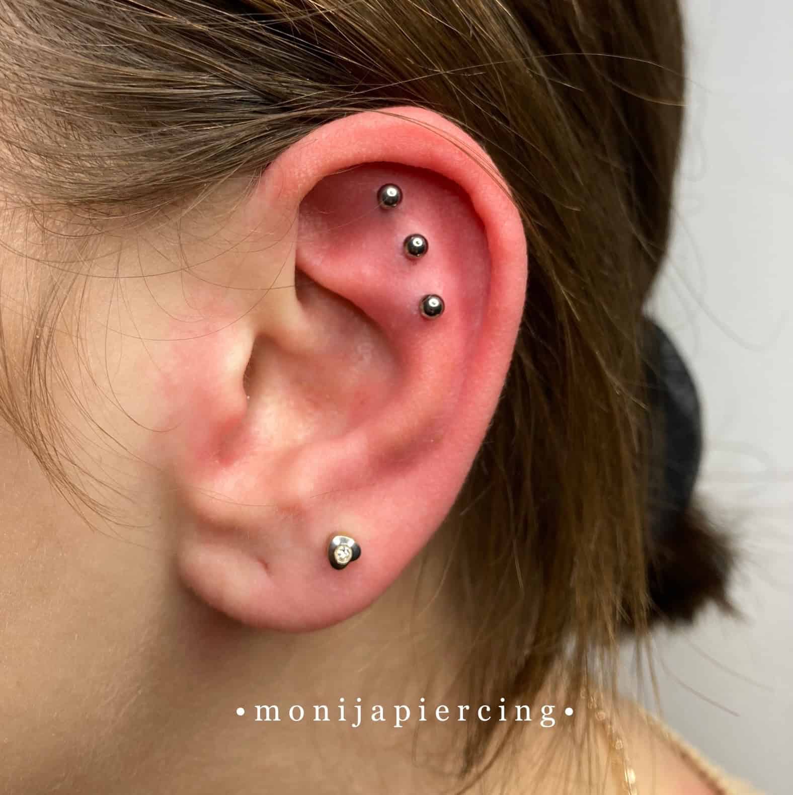 7sinstattoo.pl Zbliżenie ucha z czterema kolczykami. Trzy małe kolczyki kulkowe są ustawione pionowo wzdłuż górnej chrząstki, a pojedynczy kolczyk z przezroczystym klejnotem zdobi płatek ucha. Włosy są elegancko odgarnięte do tyłu, podkreślając kolczyki w miejscu, które można zidentyfikować jako najlepsze studio tatuażu Poznań. Tło pozostaje neutralne. Tekst: "@monijapiercing 
Nazwa produktu: IMG_0859. Najlepsze studio tatuażu Seven Sins Tattoo