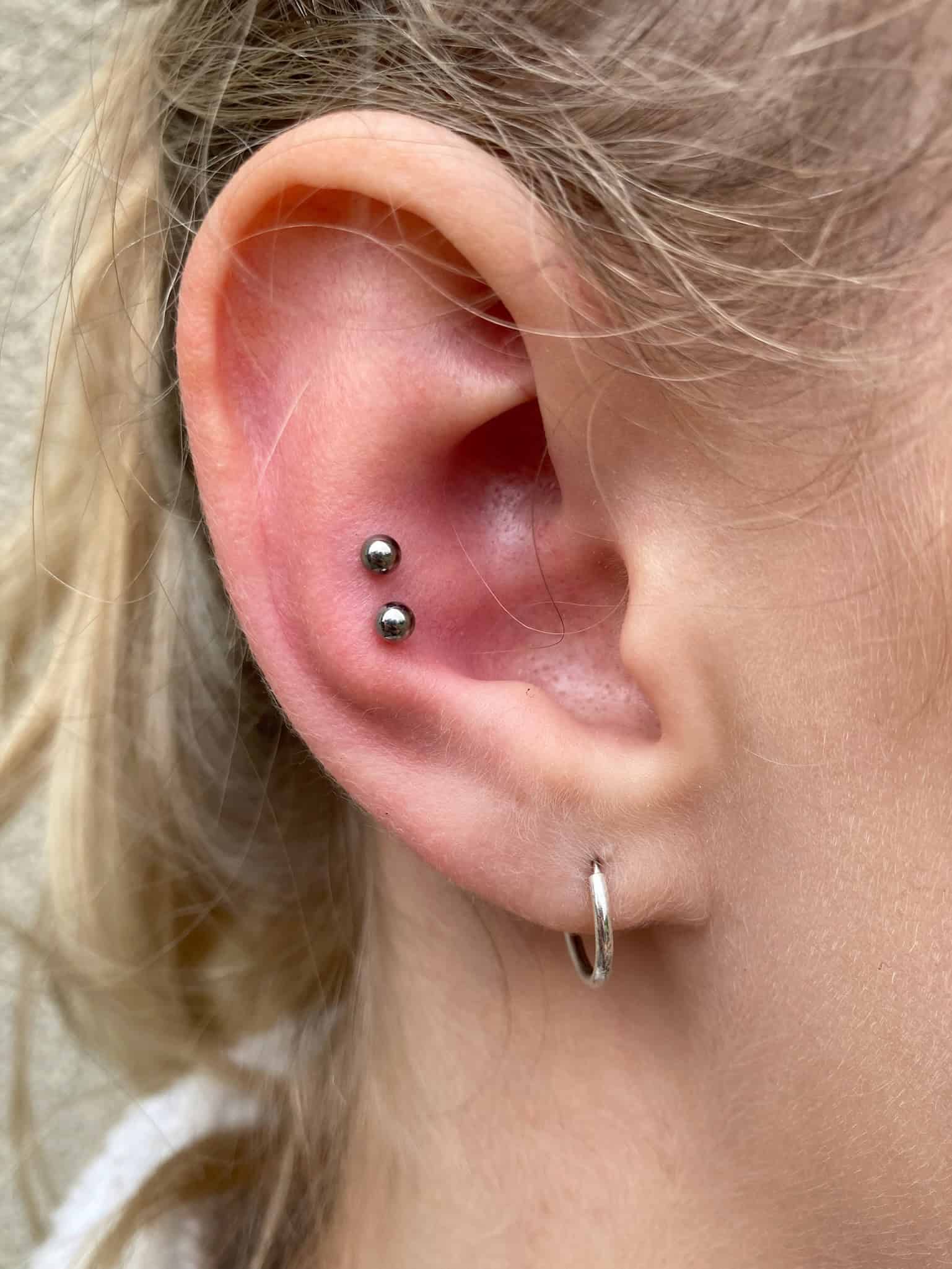 7sinstattoo.pl Zdjęcie z bliska zatytułowane IMG_1041 (1), przedstawiające ucho ozdobione dwoma srebrnymi kolczykami na górnej chrząstce i srebrnym kolczykiem-kółkiem na płatku ucha. Osoba ta ma jasnobrązowe włosy ułożone luźno na neutralnej beżowej ścianie, prawdopodobnie sfotografowane w studio tatuażu Poznań. Najlepsze studio tatuażu Seven Sins Tattoo