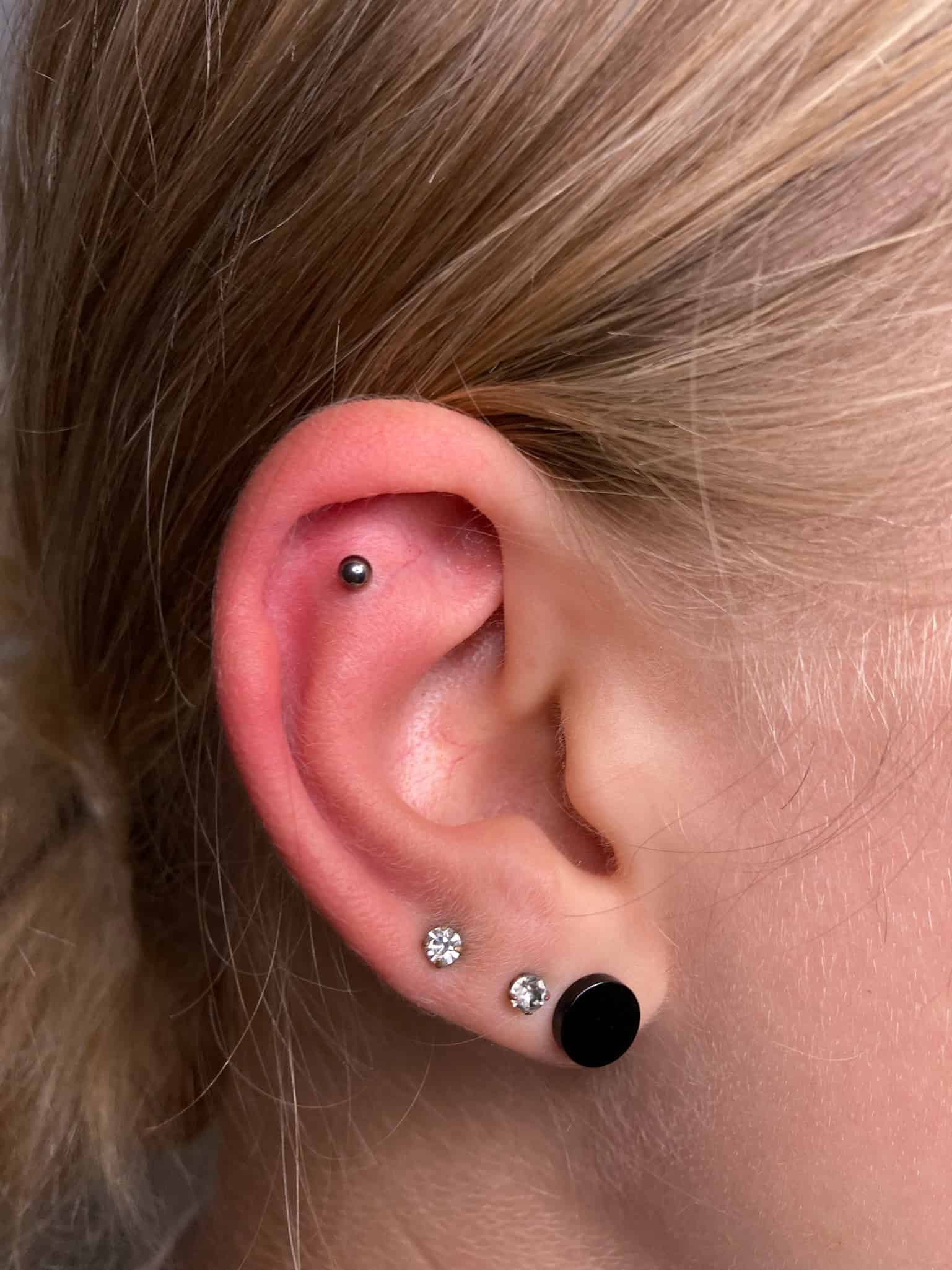 7sinstattoo.pl Zbliżenie ucha osoby z wieloma piercingami ze studia tatuażu Poznań, z Product IMG_1418. Ucho prezentuje mały srebrny kolczyk na górnej chrząstce, kolczyk z diamentem na środkowym płacie i czarny okrągły kolczyk na dolnym płacie. Tło jest rozmyte. Osoba ma jasne włosy. Najlepsze studio tatuażu Seven Sins Tattoo