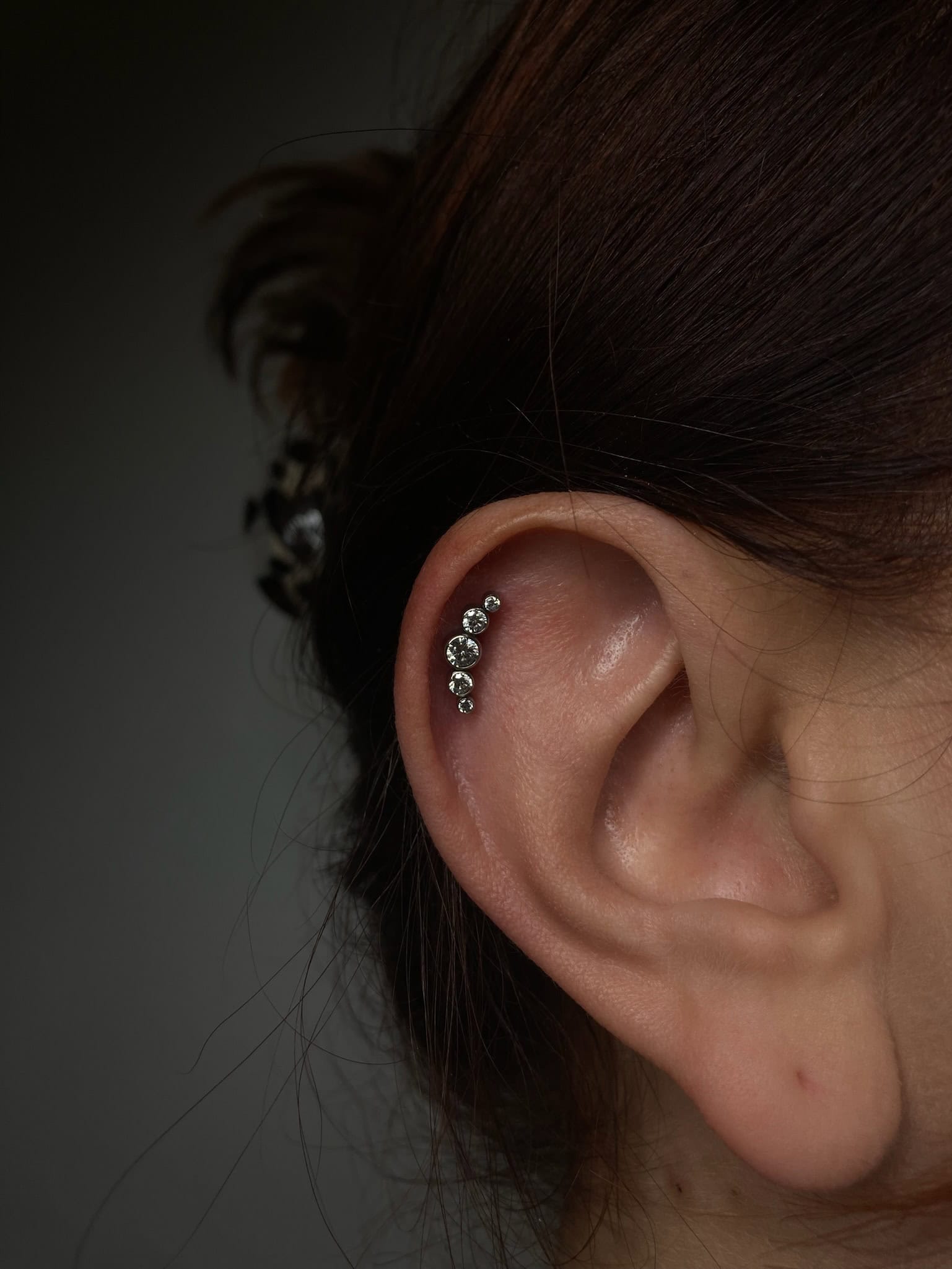 7sinstattoo.pl Zbliżenie na ucho osoby z przekłuciem chrząstki helix i rzędem pięciu małych, błyszczących kolczyków. Luźne pasma brązowych włosów oprawiają ucho, a tło stanowi ciemny, rozmyty gradient — mistrzowsko uchwycony w studio tatuażu Poznań w IMG_1546. Najlepsze studio tatuażu Seven Sins Tattoo