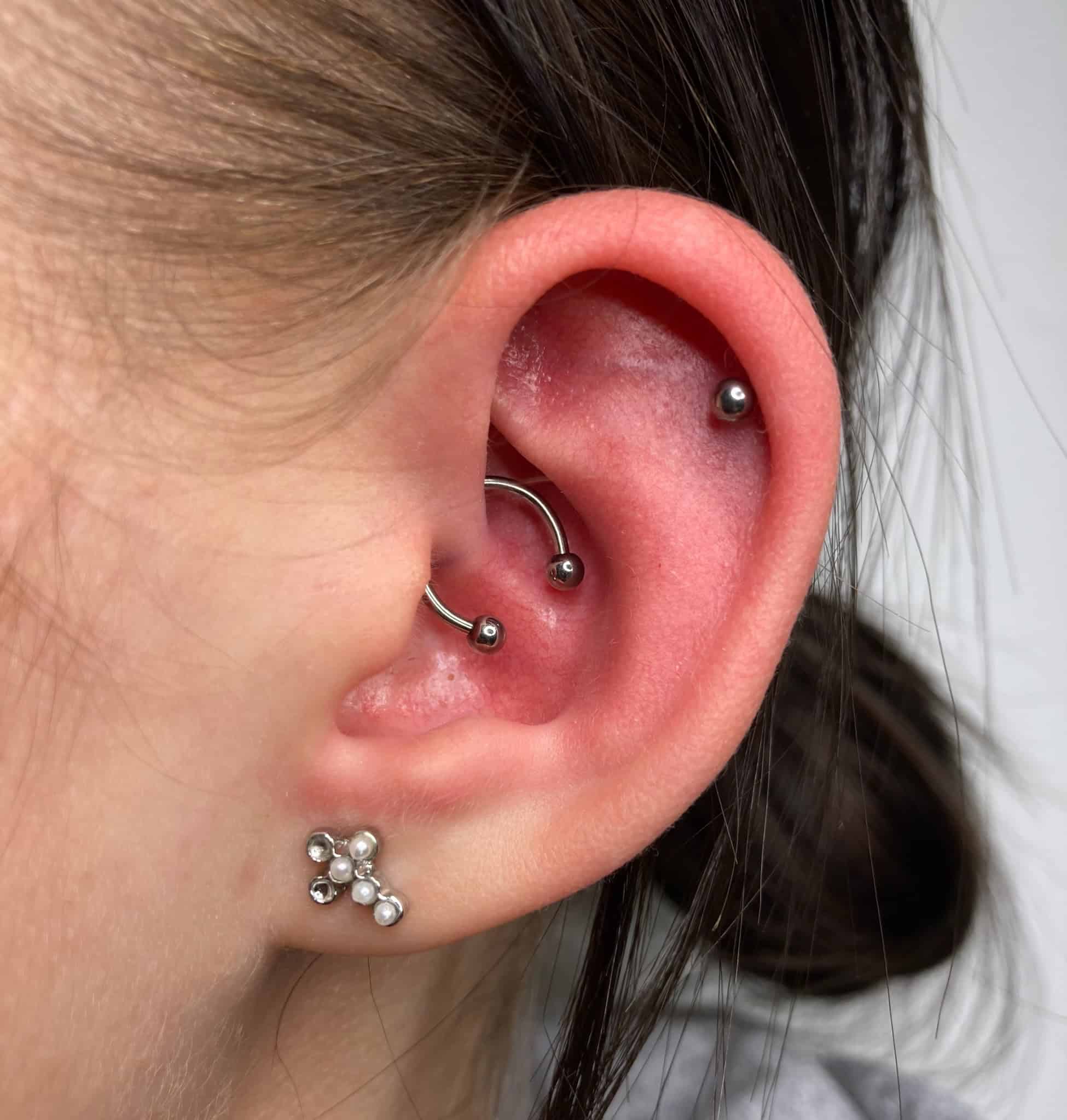 7sinstattoo.pl Na pierwszym planie widoczne jest ucho osoby ozdobione wieloma piercingami, z zakrzywionym sztangą w górnej chrząstce i małym kolczykiem w wewnętrznej muszli. Płatek ucha prezentuje elegancki kolczyk w kształcie kwiatu z płatkami pereł. Neutralne tło sugeruje, że zdjęcie zostało zrobione w najlepszym poznańskim studiu piercingu i salonie tatuażu pod nazwą produktu IMG_1633. Najlepsze studio tatuażu Seven Sins Tattoo