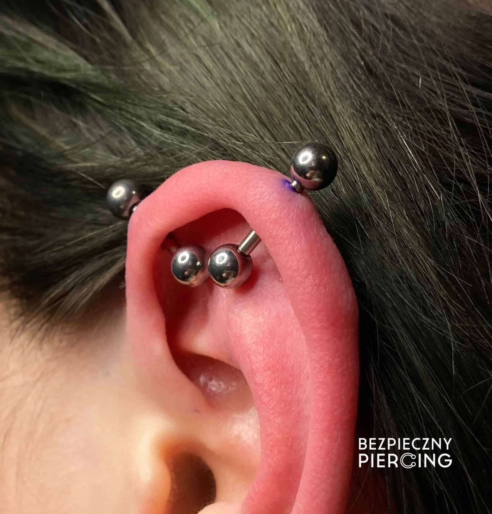 7sinstattoo.pl Zbliżenie ucha ozdobionego wieloma piercingami zaprezentowanymi w IMG_2003. Ucho ma trzy ułożone kolczyka barbell wzdłuż zewnętrznej krawędzi, co świadczy o kunszcie studia tatuażu Poznań. Piercingi wydają się być wykonane profesjonalnie. W prawym dolnym rogu napisano białym tekstem „BEZPIECZNY PIERCING”. Najlepsze studio tatuażu Seven Sins Tattoo