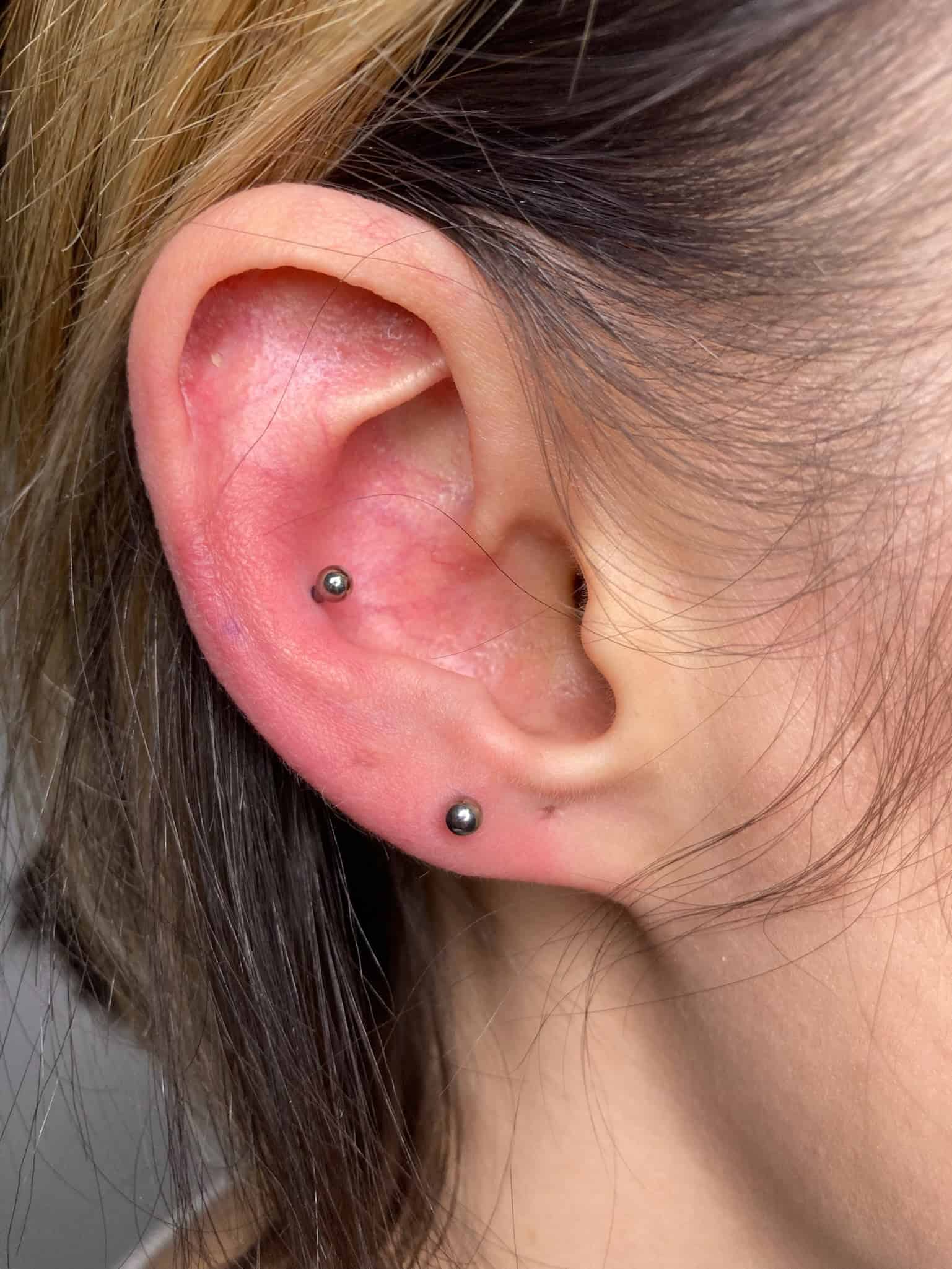 7sinstattoo.pl Zbliżenie ucha osoby z dwoma piercingami: jednym na górnej wewnętrznej chrząstce i jednym na dolnym płatku ucha, każdy z małymi, ciemnymi metalowymi ćwiekami. Jasnobrązowe włosy osoby są częściowo widoczne w ramce. Odkryj wyjątkowe style w studio tatuaży Poznań, aby uzyskać kompletny wygląd z IMG_2107! Najlepsze studio tatuażu Seven Sins Tattoo