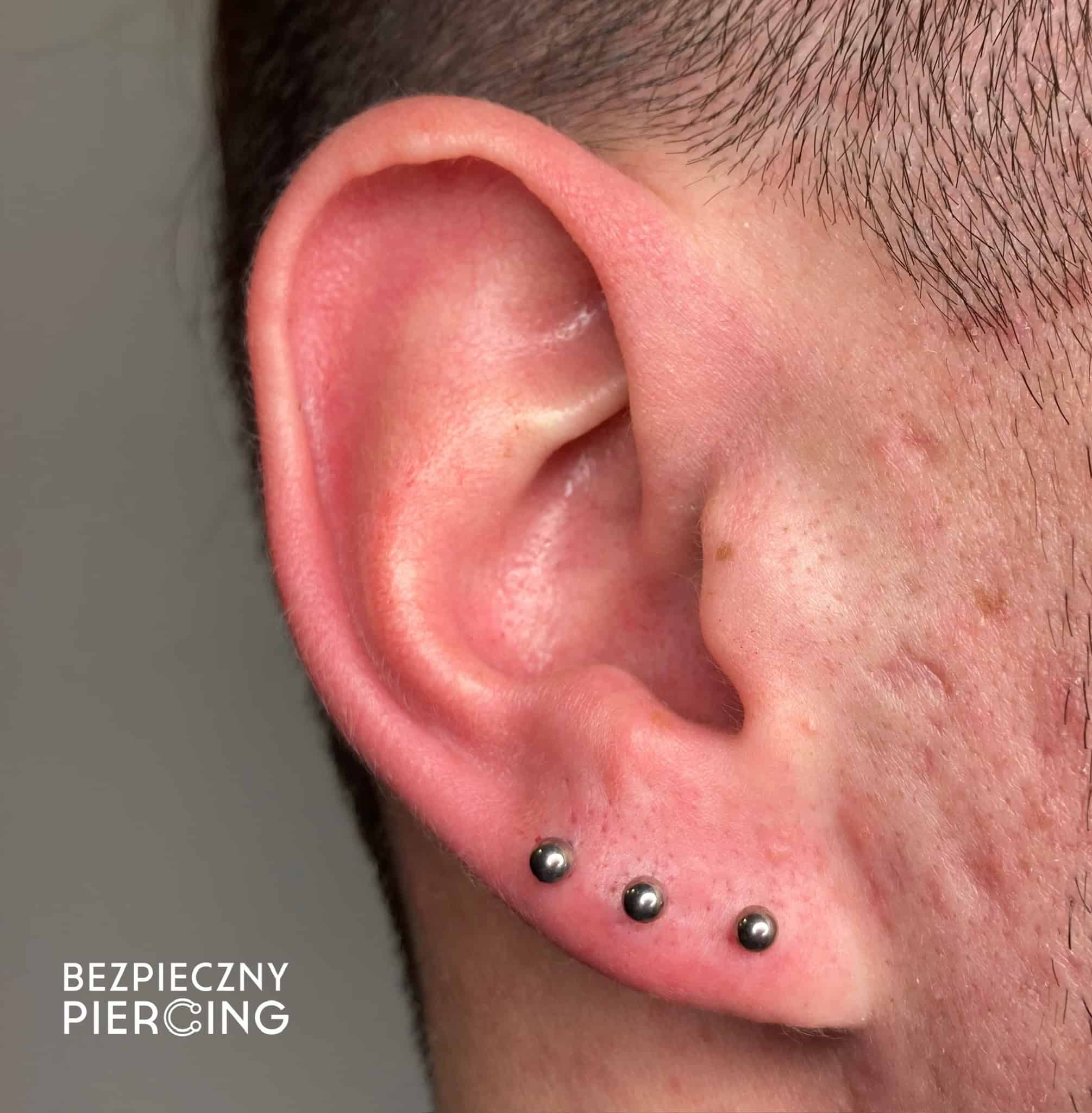 7sinstattoo.pl Zbliżenie ucha osoby z trzema srebrnymi kulkami w płatku. Tło jest neutralne, a w lewym dolnym rogu widoczny jest tekst „BEZPIECZNY PIERCING”. Skóra w pobliżu piercingu jest lekko różowa, co sugeruje niedawne przekłucie w studio tatuaży Poznań. Nazwa produktu: IMG_2232. Najlepsze studio tatuażu Seven Sins Tattoo
