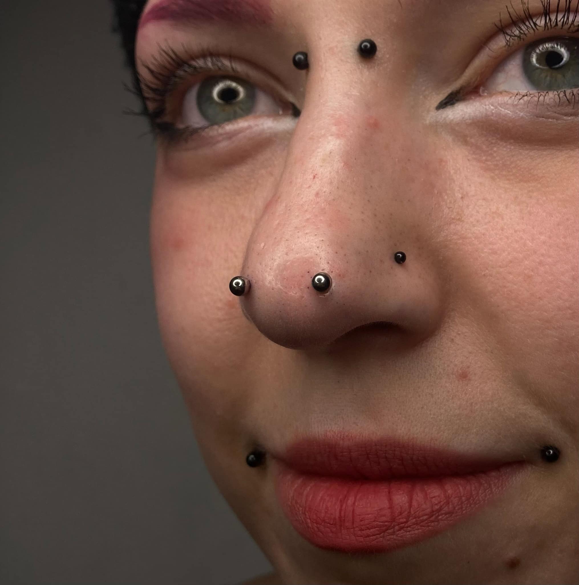 7sinstattoo.pl Zbliżenie twarzy osoby z wieloma piercingami twarzy, w tym dwoma kolczykami w nosie, kolczykiem w przegrodzie nosowej i parą piercingów wargi dolnej. Osoba ma różowy makijaż oczu, skrzydlatą kreskę na powiece i jasnoczerwoną szminkę. Odkryj ten styl na naszym wyróżnionym zdjęciu: IMG_2240 w najlepsze studio tatuażu Poznań. Najlepsze studio tatuażu Seven Sins Tattoo