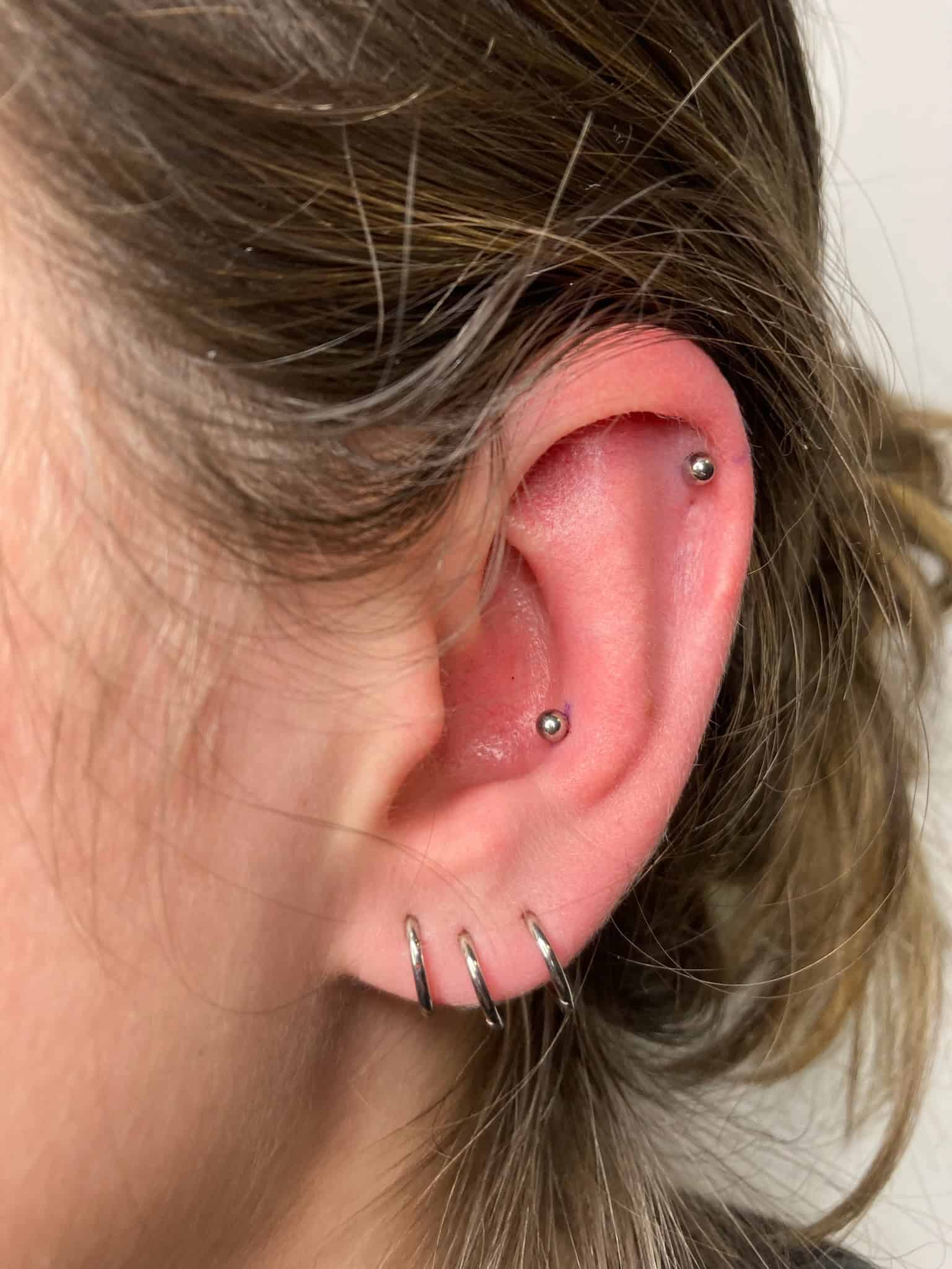 7sinstattoo.pl Zbliżenie ucha osoby z trzema piercingami stylizowanymi za pomocą IMG_2278. Dwa małe kolczyki w kształcie kółek zdobią dolny płatek ucha, a pojedynczy kolczyk zdobi helisę (górną chrząstkę ucha). Jasnobrązowe włosy osoby są częściowo odgarnięte do tyłu, co zapewnia wypolerowany wygląd przypominający kunszt studio tatuaży Poznań. Najlepsze studio tatuażu Seven Sins Tattoo