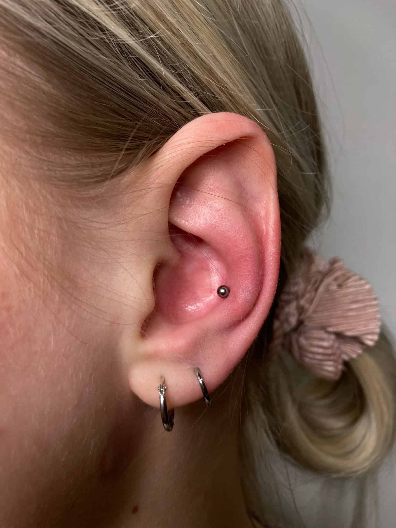 7sinstattoo.pl Zdjęcie produktu IMG_2753 przedstawia zbliżenie ucha osoby ozdobionego piercingiem muszli, stylowo zaakcentowanym małym srebrnym kolczykiem kulkowym. Osoba ta nosi również dwa kolczyki w kształcie kółek w płatku ucha, a jej blond włosy są starannie związane za pomocą gumki do włosów z teksturą, co doskonale prezentuje wykwintne kunszt, jakiego można oczekiwać od najlepszego studia tatuażu w Poznaniu. Najlepsze studio tatuażu Seven Sins Tattoo