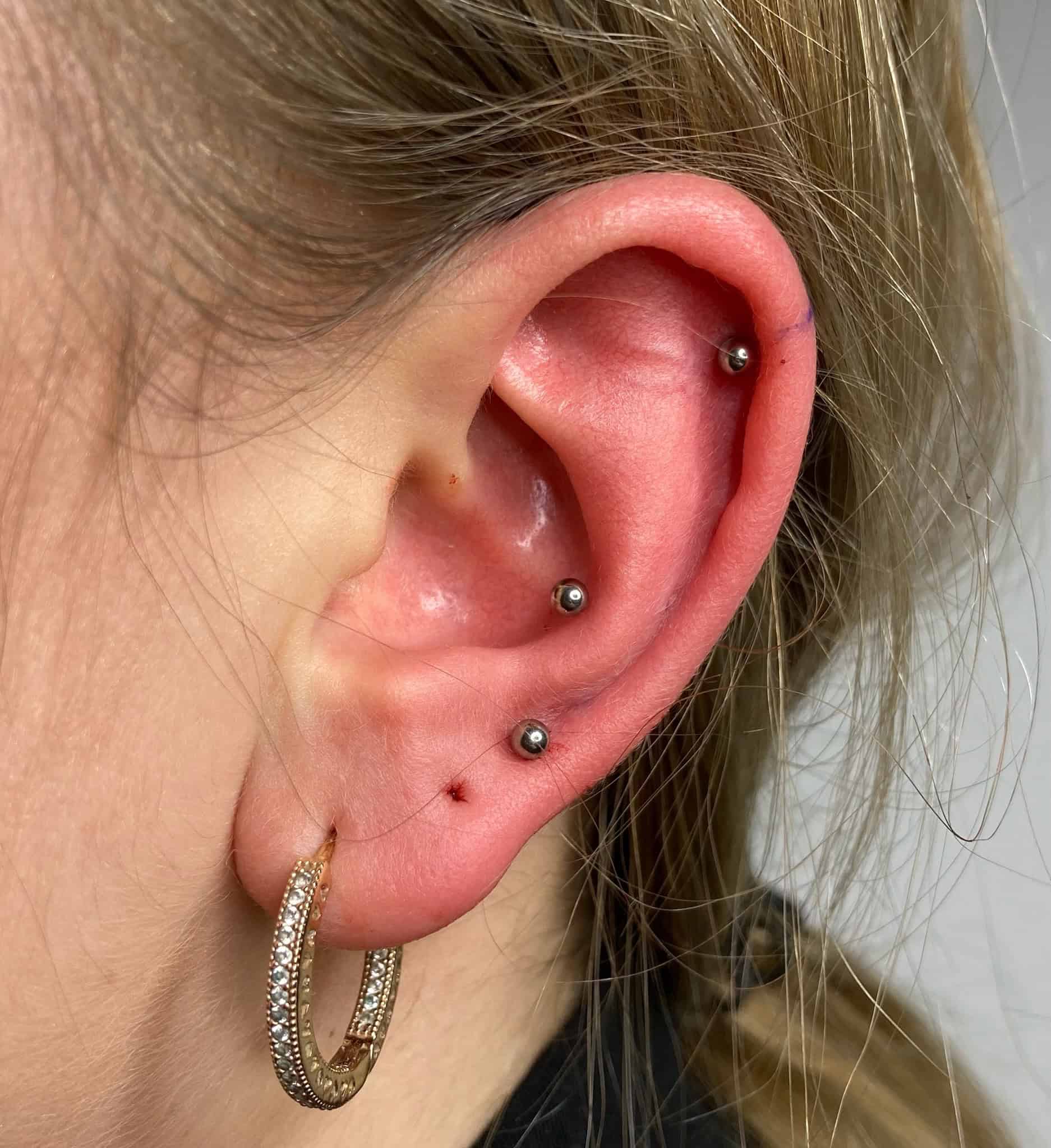 7sinstattoo.pl Zdjęcie z bliska, zidentyfikowane jako IMG_2797, przedstawia ucho osoby ozdobione kilkoma srebrnymi kolczykami. Ucho ma trzy ćwieki na chrząstce i jeden na płatku, uzupełnione złotym kolczykiem w kształcie koła z kryształkami. Jasne włosy osoby są związane z tyłu i częściowo widoczne na zdjęciu, które najwyraźniej zostało zrobione w studio tatuażu Poznań. Najlepsze studio tatuażu Seven Sins Tattoo