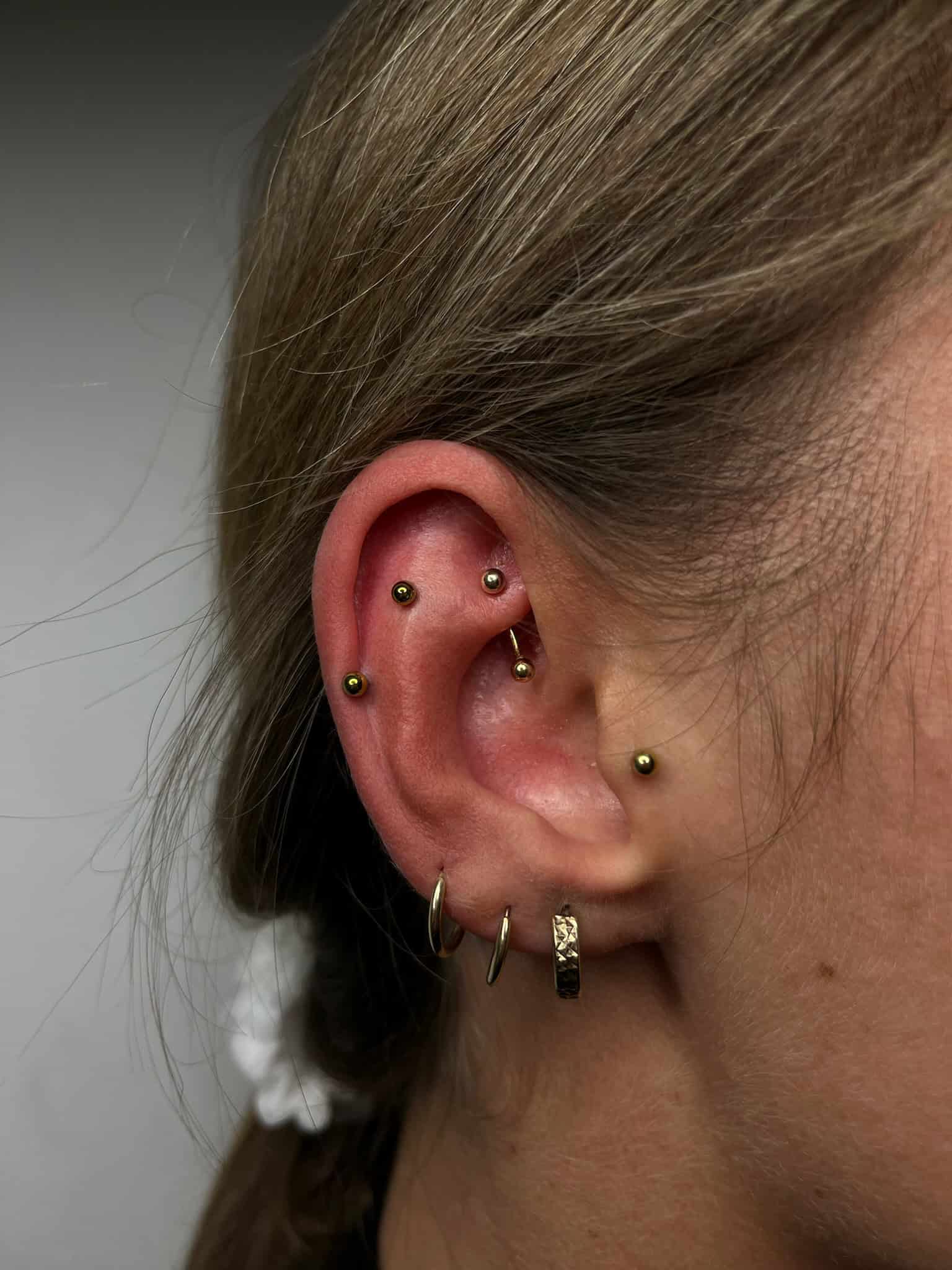 7sinstattoo.pl Zbliżenie z IMG_3037 pokazuje ucho ozdobione wieloma piercingami, idealne dla osób odwiedzających najlepsze studio tatuażu w Poznaniu. Ucho ma kilka złotych kolczyków wzdłuż chrząstki i kilka złotych kółek na płatku ucha. Włosy osoby są ściągnięte do tyłu, zaakcentowane białym ozdobą do włosów. Najlepsze studio tatuażu Seven Sins Tattoo