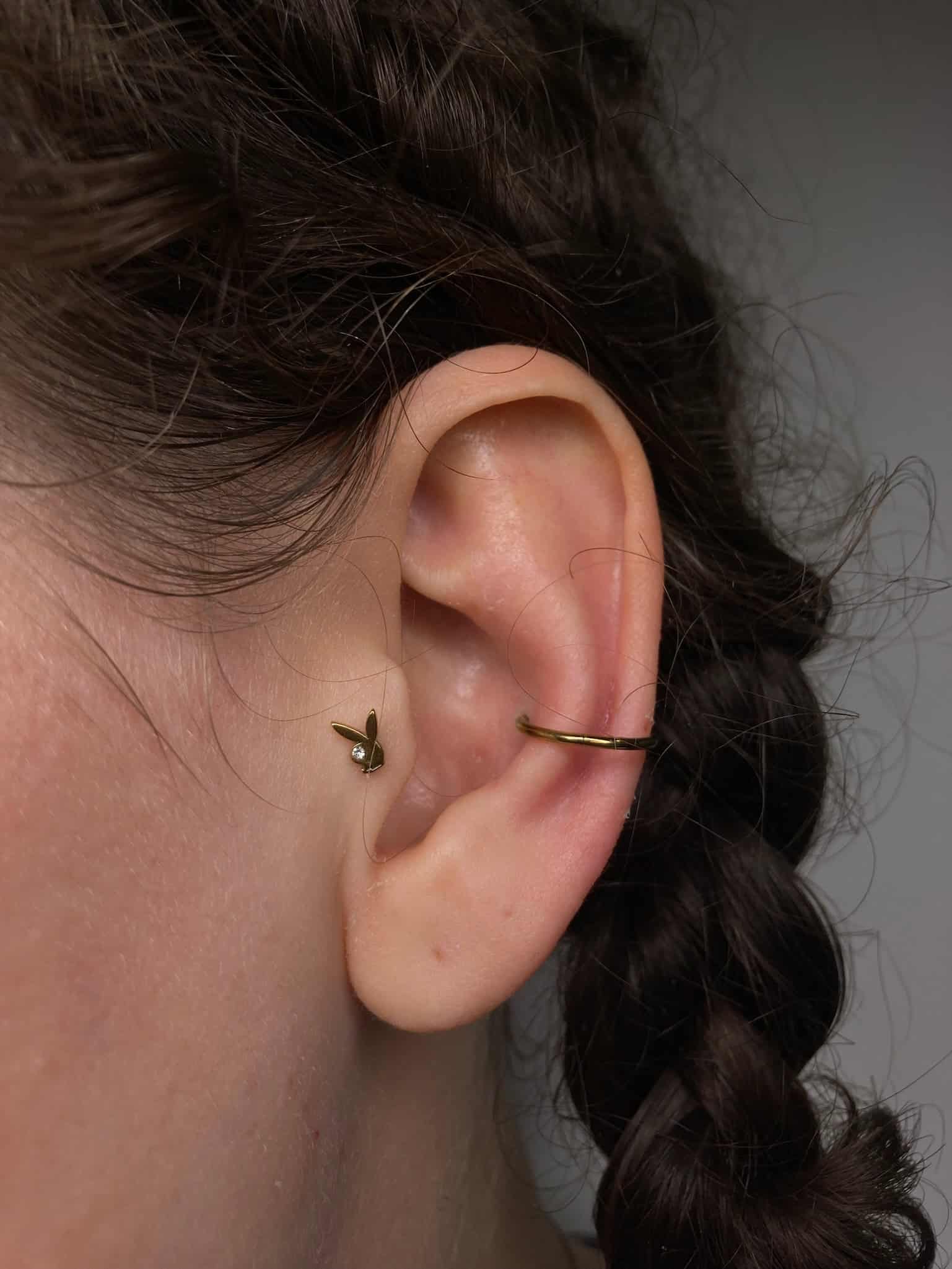 7sinstattoo.pl Zbliżenie ucha osoby ozdobionego dwoma kolczykami IMG_3073 z najlepszego studia tatuażu Poznań. Ucho ma mały kolczyk w kształcie liścia na górnym płatku i prosty kolczyk w kształcie kółka na helisie. Warkocz włosów osoby opada obok ucha. Najlepsze studio tatuażu Seven Sins Tattoo