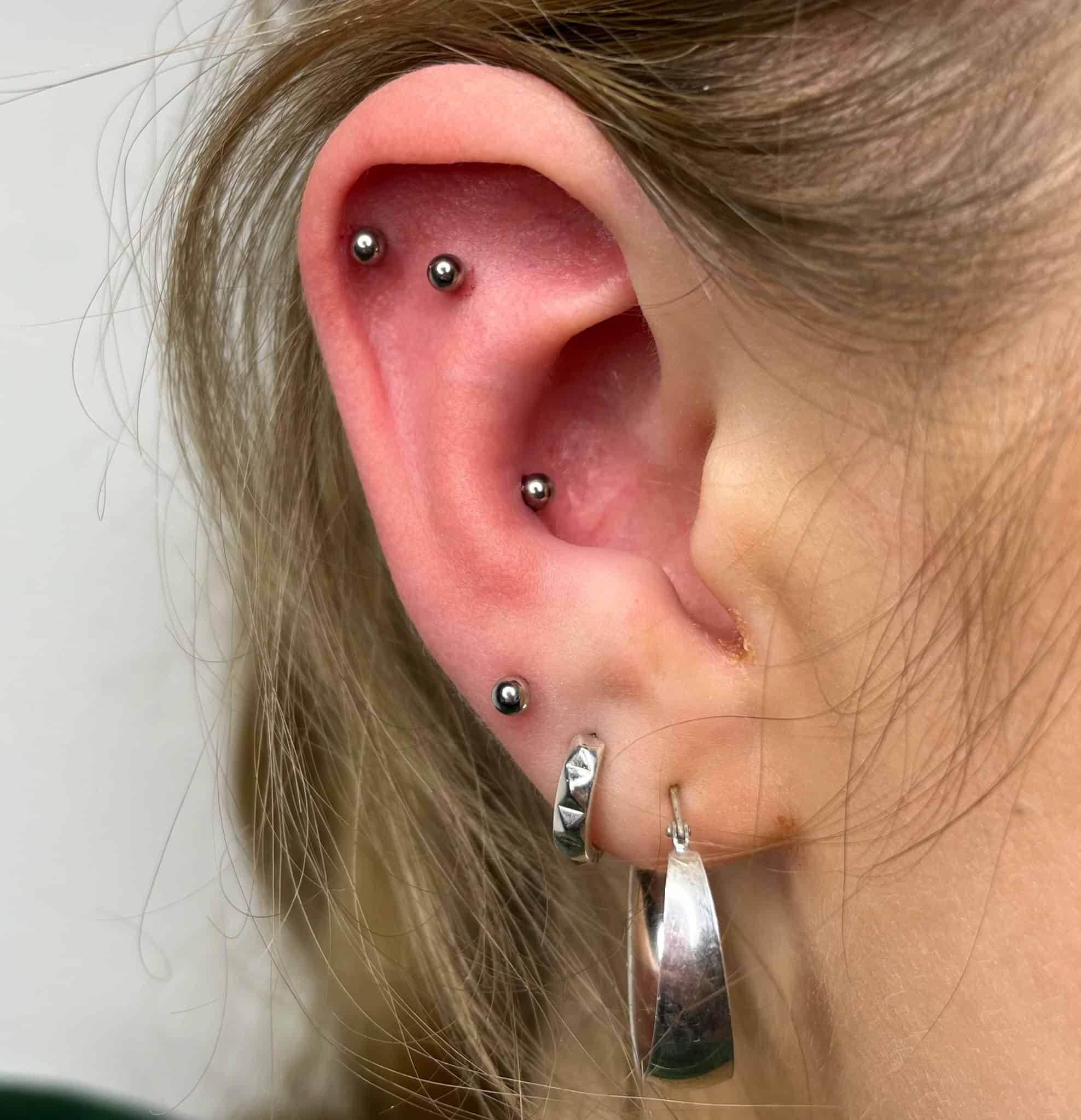 7sinstattoo.pl Opis produktu dla IMG_3100: 
Zbliżenie lewego ucha osoby ozdobionego wieloma piercingami, zrobione w studio tatuażu Poznań. Ucho ma dwa małe, srebrne ćwieki na górnej części, mały kółko obok wewnętrznego ucha i dwa srebrne kolczyki w kształcie kółek na dolnym płatku — jeden gładki i jeden teksturowany. Najlepsze studio tatuażu Seven Sins Tattoo