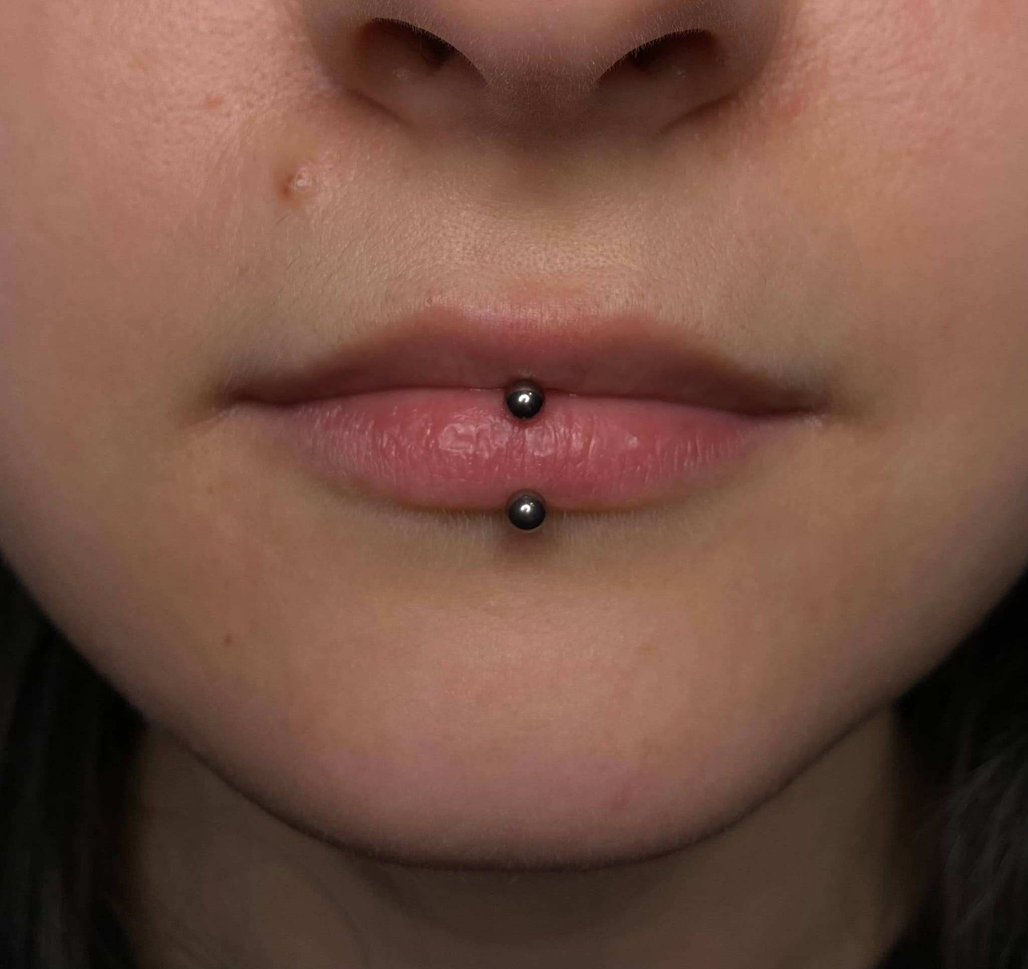 7sinstattoo.pl Zbliżenie ust osoby z pionowym piercingiem wargi. Piercing składa się z dwóch małych, czarnych kulek — jednej na dolnej wardze, a drugiej poniżej — połączonych prętem. Uchwycony w IMG_3158 w najlepsze studio tatuażu Poznań, tło nie jest widoczne, skupiając się tylko na dolnej części twarzy. Najlepsze studio tatuażu Seven Sins Tattoo