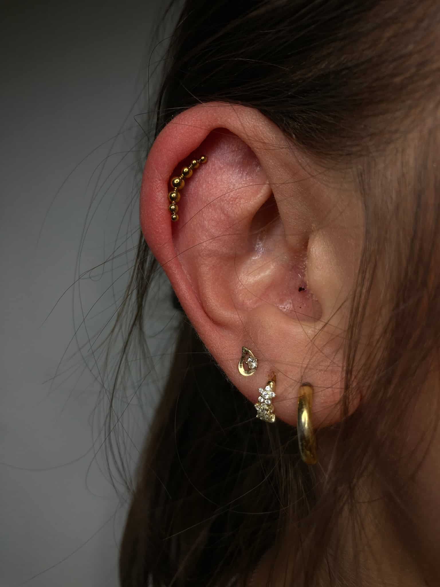 7sinstattoo.pl Zbliżenie na ludzkie ucho ozdobione wieloma piercingami, prezentujące różne kolczyki, w tym zakrzywiony rząd małych złotych koralików wzdłuż helisy, gwiazdę, półksiężyc, mały sztyft i prosty złoty kółko na płatku uchwycony idealnie na zdjęciu IMG_3569 autorstwa studia tatuażu Poznań. Najlepsze studio tatuażu Seven Sins Tattoo