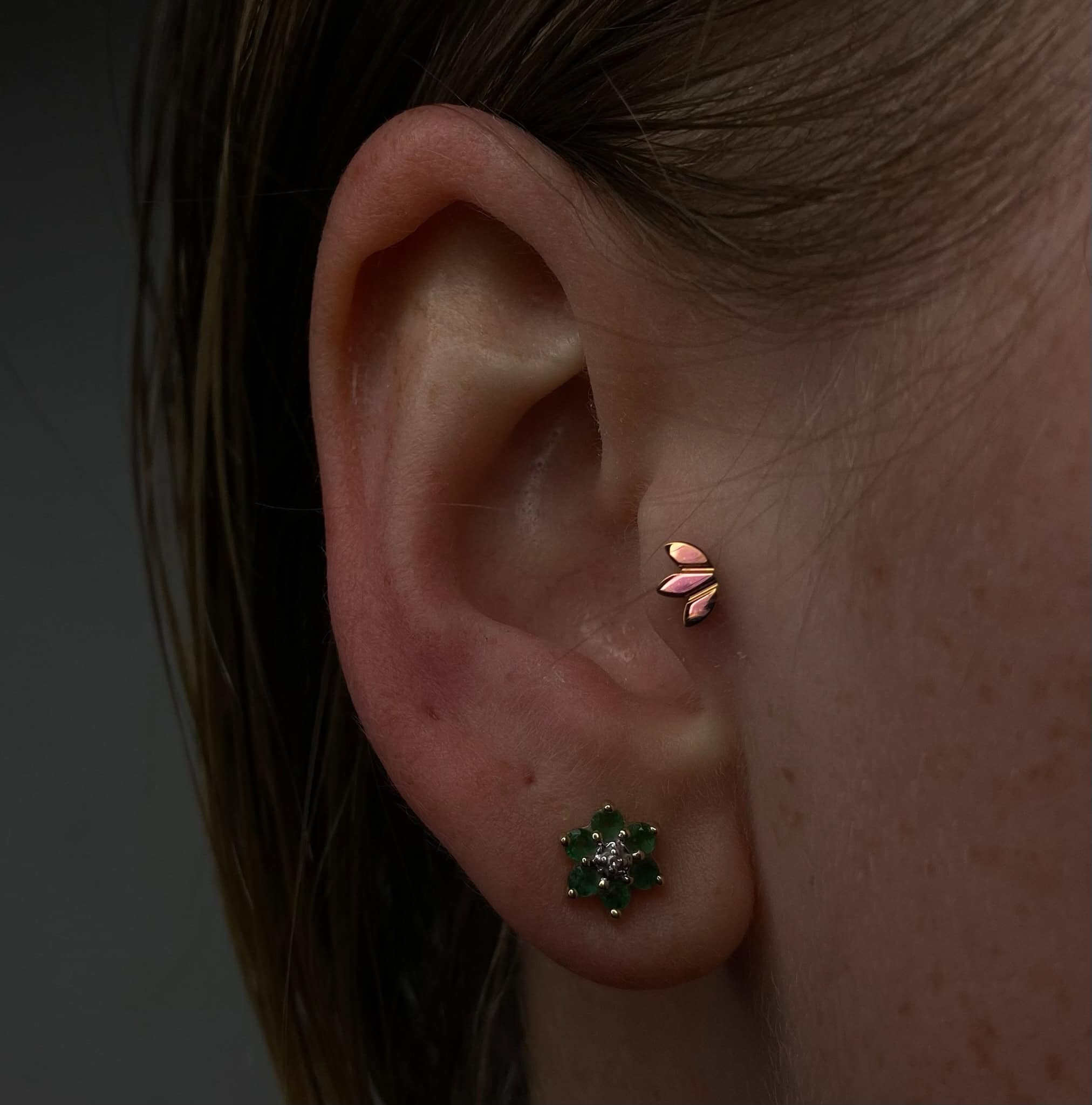 7sinstattoo.pl Zbliżenie ucha osoby, z dwoma kolczykami, wyróżnia się. Dolny kolczyk ma zielony kamień szlachetny w kwiatowym wzorze, podczas gdy górny kolczyk jest wykonany z różowego złota z minimalistycznym motywem trzech liści. Idealne akcesoria dla kogoś, kto planuje odwiedzić najlepsze studio tatuażu w Poznaniu, pięknie uchwycone na zdjęciu produktu IMG_3584. Najlepsze studio tatuażu Seven Sins Tattoo