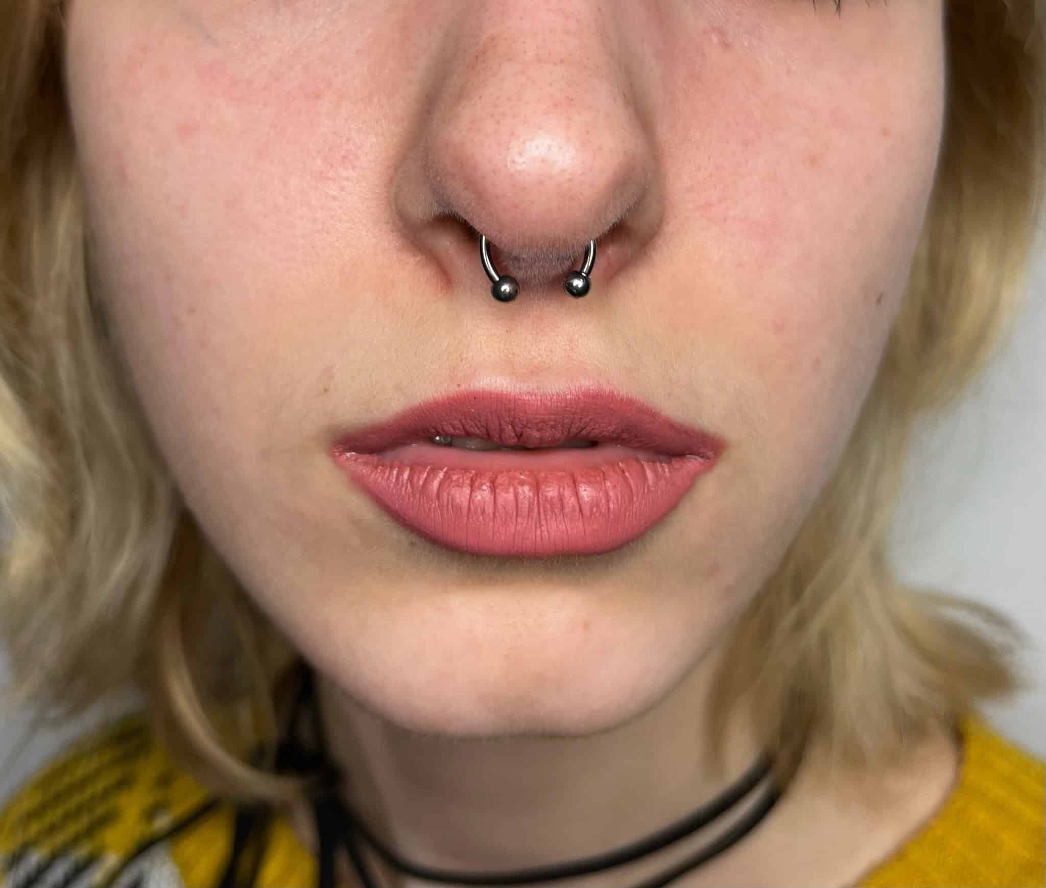 7sinstattoo.pl Zbliżenie twarzy osoby, skupiające się na nosie i ustach. Osoba ma piercing przegrody nosowej z kółkiem w kształcie podkowy i nosi jasnoróżową szminkę. W tle widoczne są również blond włosy i część żółtego ubrania. Zrobione w Studio Tatuaże Poznań, znanym ze swojego artystycznego stylu. Nazwa produktu: IMG_4201. Najlepsze studio tatuażu Seven Sins Tattoo