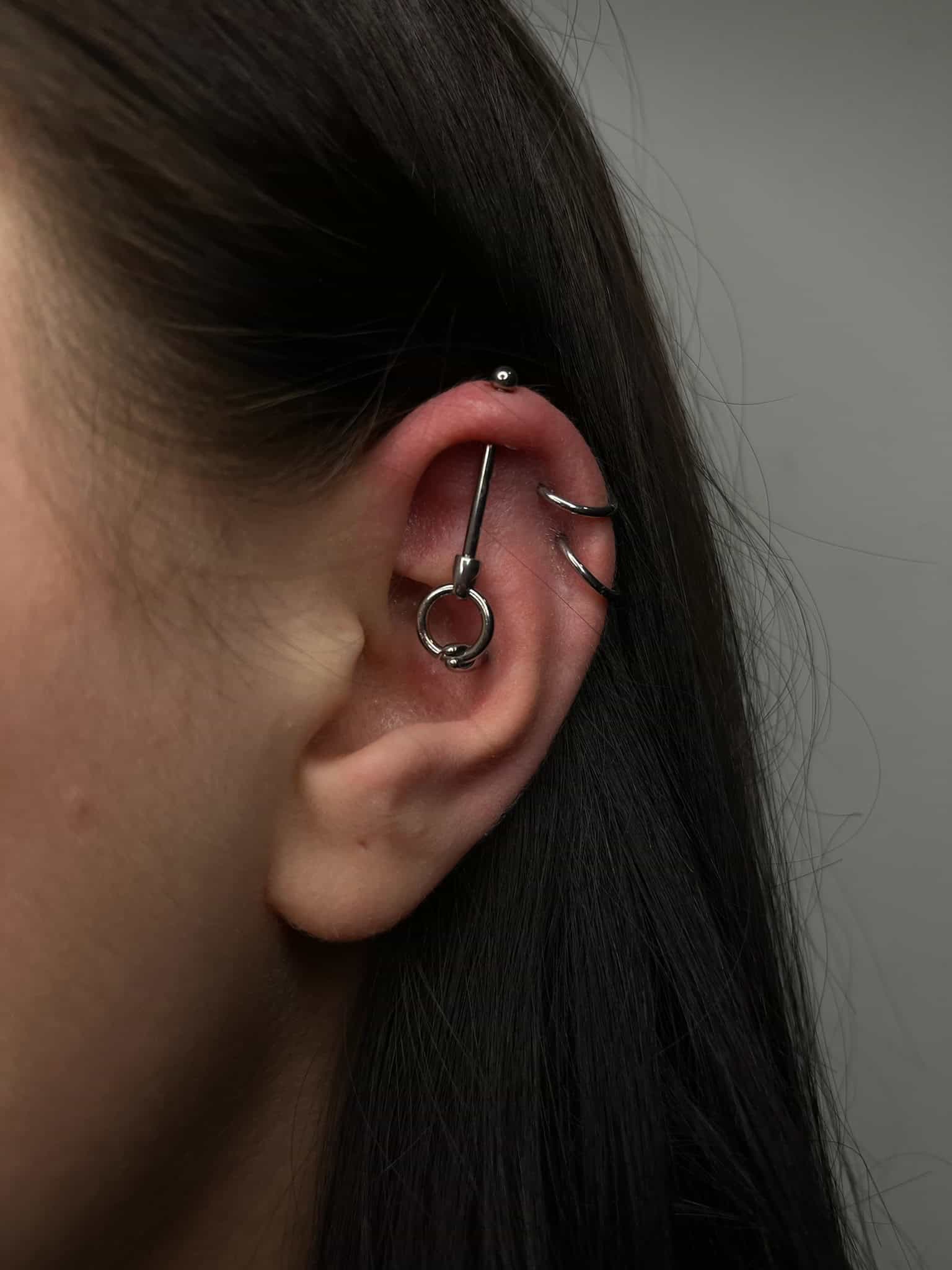 7sinstattoo.pl Zbliżenie ucha ozdobionego różnymi piercingami, w tym małym kolczykiem kulkowym przy górnej krawędzi, kolczykiem w kształcie kółka otaczającym środek i industrialnym prętem przebijającym chrząstkę. Ucho wyróżnia się na neutralnym szarym tle, co jest przykładem kunsztu studia tatuażu Poznań. Ten obraz nosi tytuł IMG_4409. Najlepsze studio tatuażu Seven Sins Tattoo