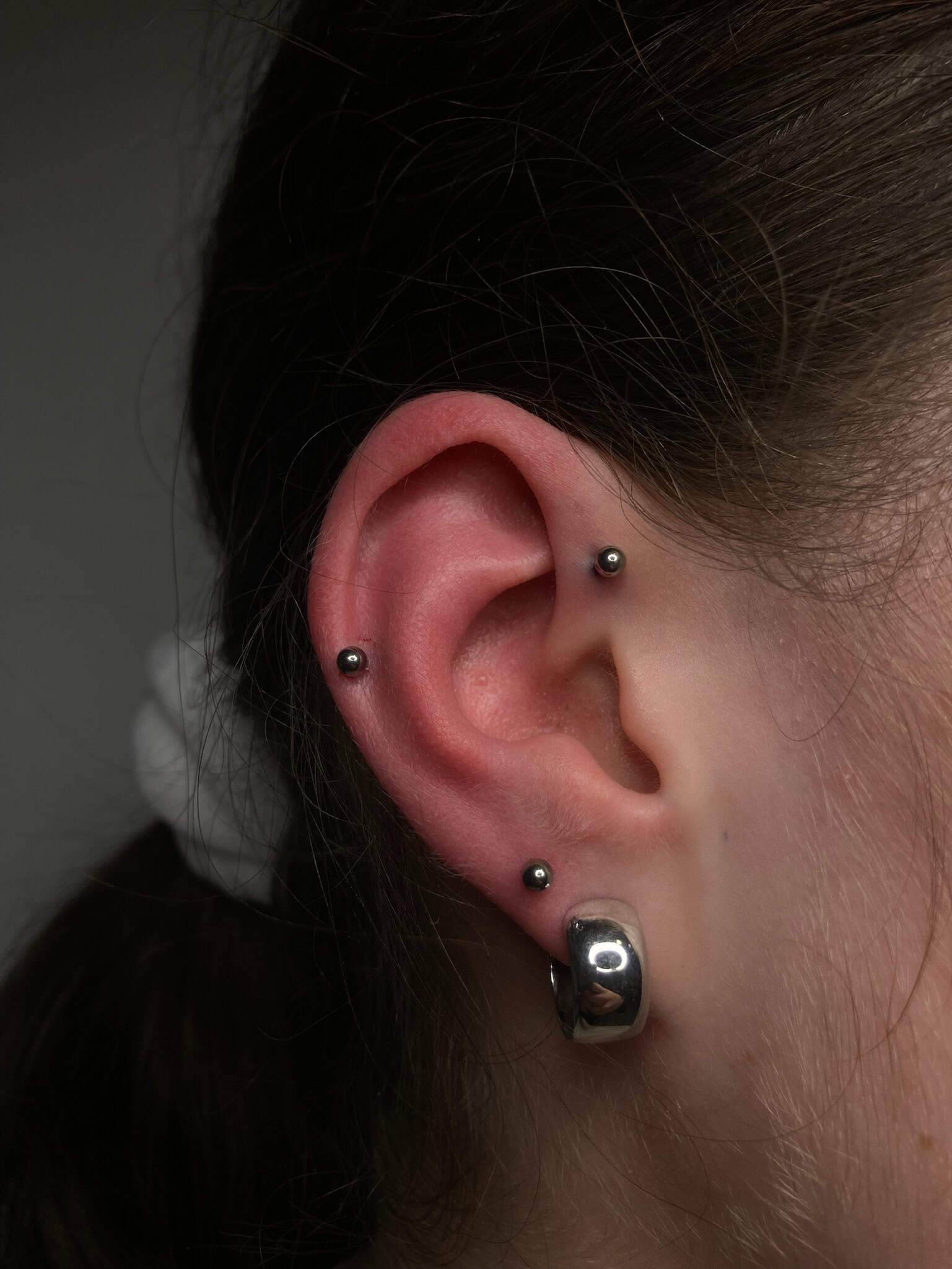 7sinstattoo.pl Zbliżenie ucha z wieloma kolczykami, zrobione w studio tatuażu Poznań. Obraz o nazwie IMG_4451 przedstawia dwa małe kolczyki umieszczone na górze i środku ucha, a także większy kolczyk-kółko na dole. Osoba ma ciemne włosy związane z tyłu, co widać po lewej stronie obrazu. Najlepsze studio tatuażu Seven Sins Tattoo