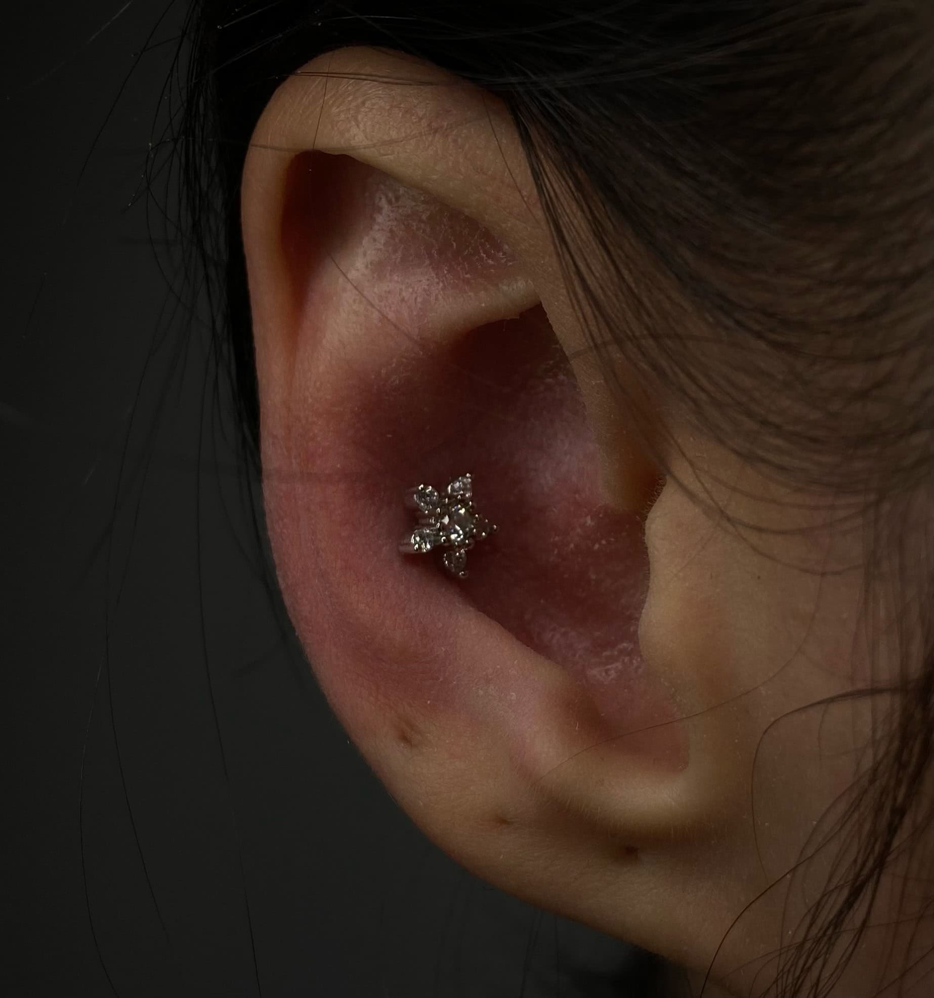 7sinstattoo.pl Zbliżenie ucha z małym, błyszczącym diamentowym piercingiem chrząstki ozdobionym wzorem w kształcie kwiatu. Ucho jest oświetlone na ciemnym tle, podkreślając misterne detale i blask IMG_4764. Pasma włosów są widoczne w pobliżu w studio tatuaży Poznań. Najlepsze studio tatuażu Seven Sins Tattoo