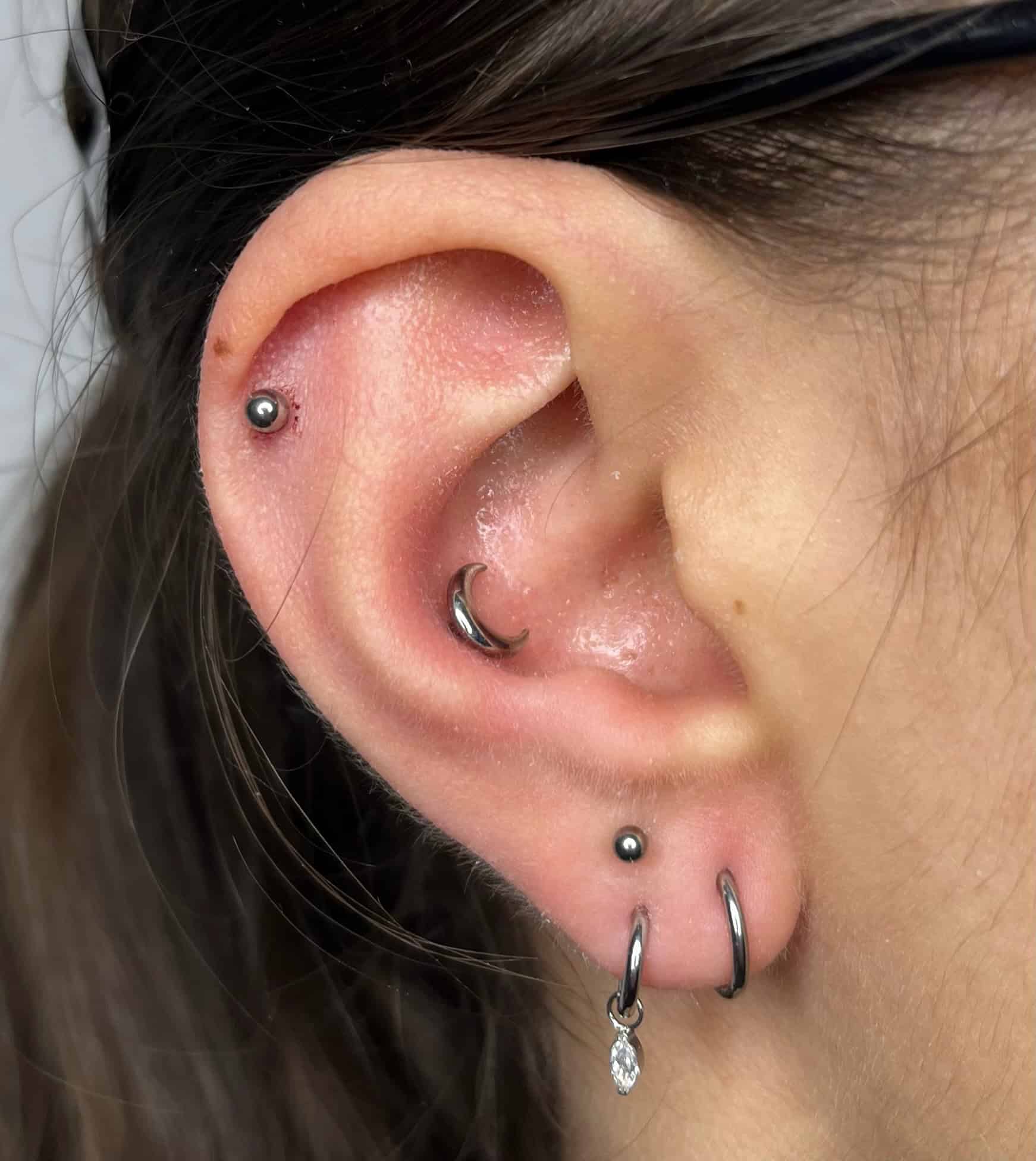 7sinstattoo.pl Na zdjęciu IMG_4797 zbliżenie ukazuje ucho osoby ozdobione wieloma srebrnymi kolczykami: małym kółkiem w płatku ucha, kolczykiem kulkowym nad nim, zakrzywionym sztangą w uchu wewnętrznym i kolczykiem w górnej chrząstce. Osoba ma ciemne włosy i nosi okulary. Odwiedź studio tatuażu Poznań, aby zobaczyć więcej inspirujących tatuaży na ciele. Najlepsze studio tatuażu Seven Sins Tattoo