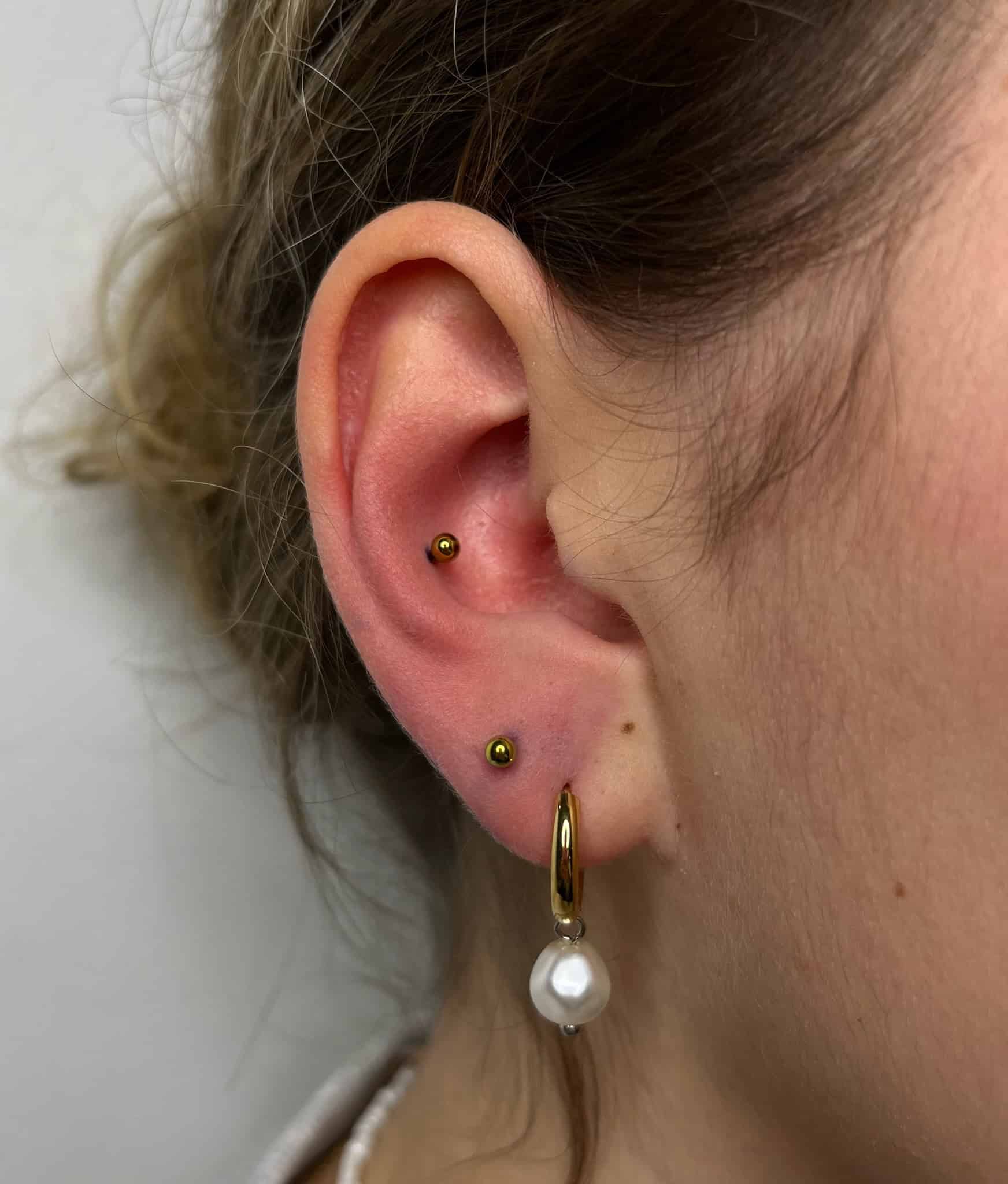 7sinstattoo.pl Nieskazitelna fotografia oznaczona jako IMG_4849 ze studia tatuażu Poznań uchwyca z bliska ucho osoby ozdobione dwoma małymi złotymi kolczykami w górnej chrząstce i większym złotym kolczykiem w kształcie koła z wiszącą perłą w płatku ucha. Włosy są starannie zaczesane do tyłu, eksponując całe ucho na prostym i neutralnym tle. Najlepsze studio tatuażu Seven Sins Tattoo