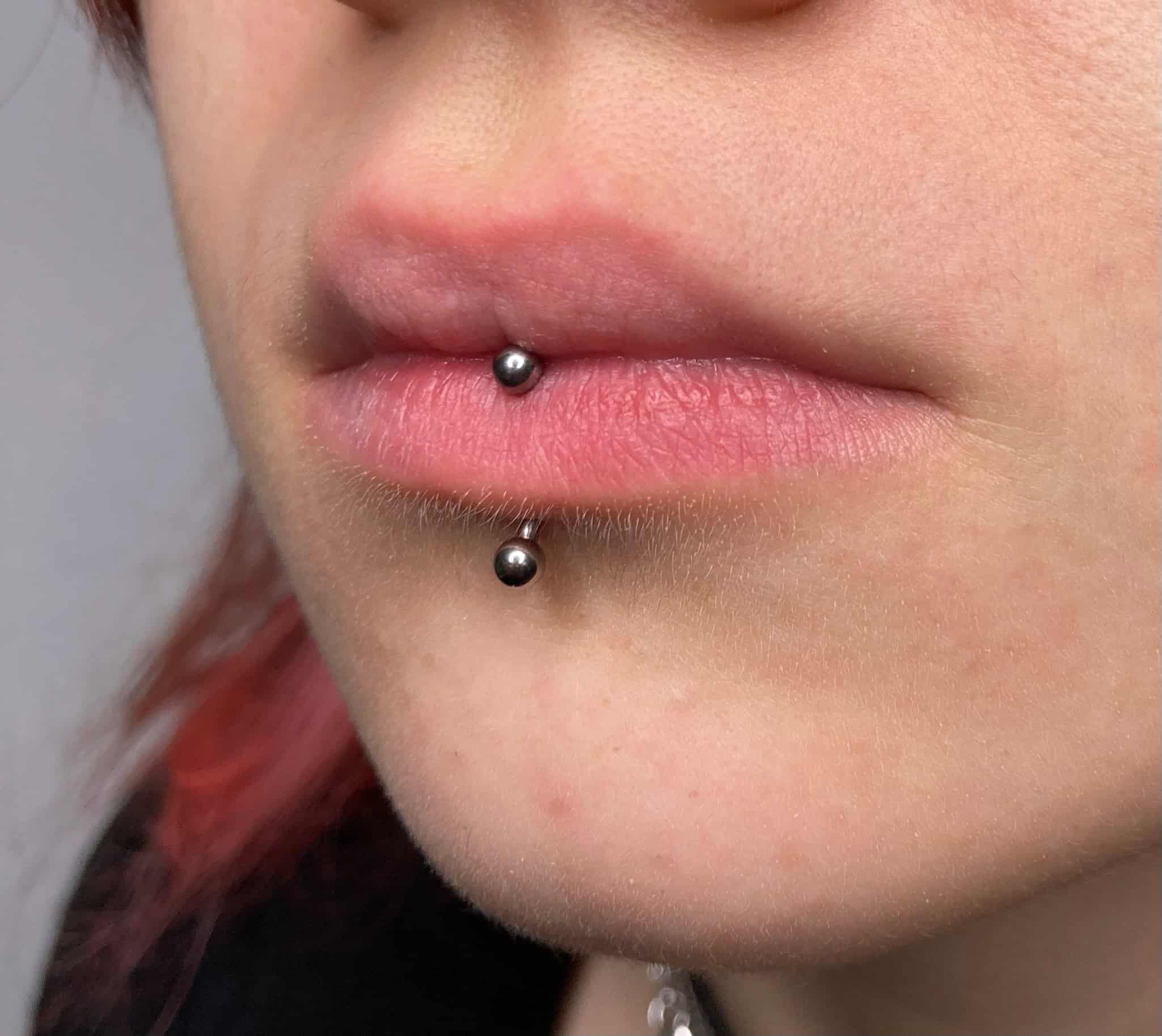 7sinstattoo.pl Zbliżenie ust osoby pokazujące podwójne pionowe piercingi wargi górnej. Górny piercing znajduje się na dole dolnej wargi, a dolny piercing tuż nad brodą. Oba piercingi mają małą, okrągłą metalową biżuterię. Zdjęcie, zidentyfikowane jako IMG_4897, zostało zrobione w renomowanym studiu tatuaży Poznań na neutralnym tle. Najlepsze studio tatuażu Seven Sins Tattoo