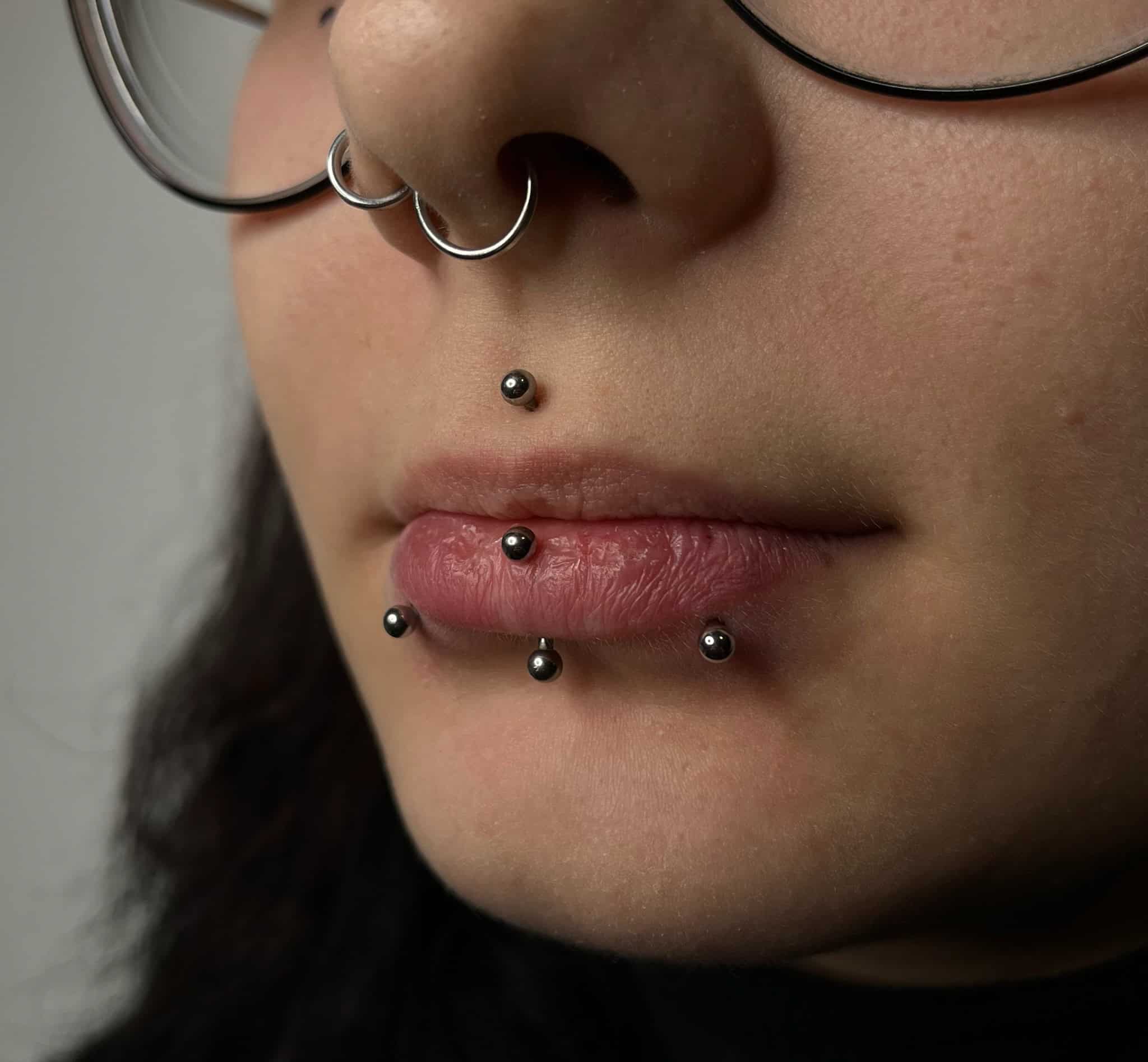 7sinstattoo.pl Zbliżenie osoby noszącej okulary, z piercingiem twarzy. Piercing obejmuje kółko w przegrodzie nosowej, kolczyk w wargę górną i poziomy rząd trzech kolczyków pod dolną wargą. Uchwycony w studio tatuażu Poznań, IMG_5168 przedstawia osobę z neutralnym wyrazem twarzy. Najlepsze studio tatuażu Seven Sins Tattoo