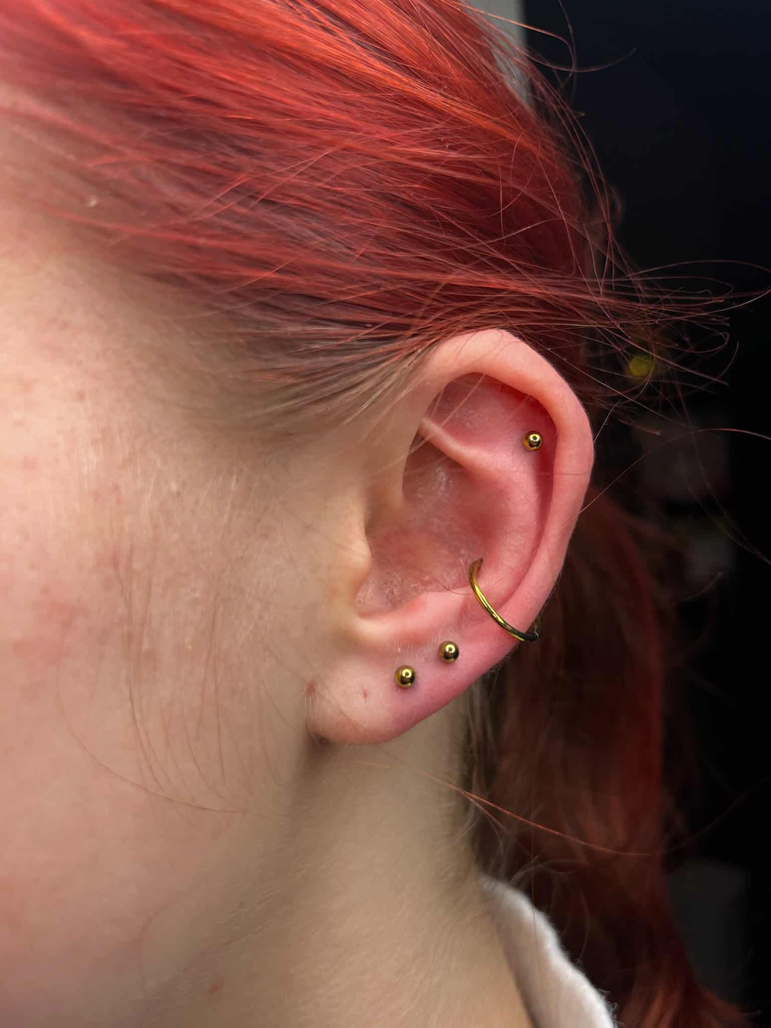 7sinstattoo.pl Zdjęcie z bliska zatytułowane IMG_5427 przedstawia ucho osoby ozdobione kilkoma złotymi kolczykami, zrobione w najlepszym studio tatuażu Poznań. Ucho ma mały złoty kolczyk u góry, złoty pierścień w połowie drogi i dwa złote kolczyki na płatku ucha. Osoba ma efektowne rude włosy. Najlepsze studio tatuażu Seven Sins Tattoo