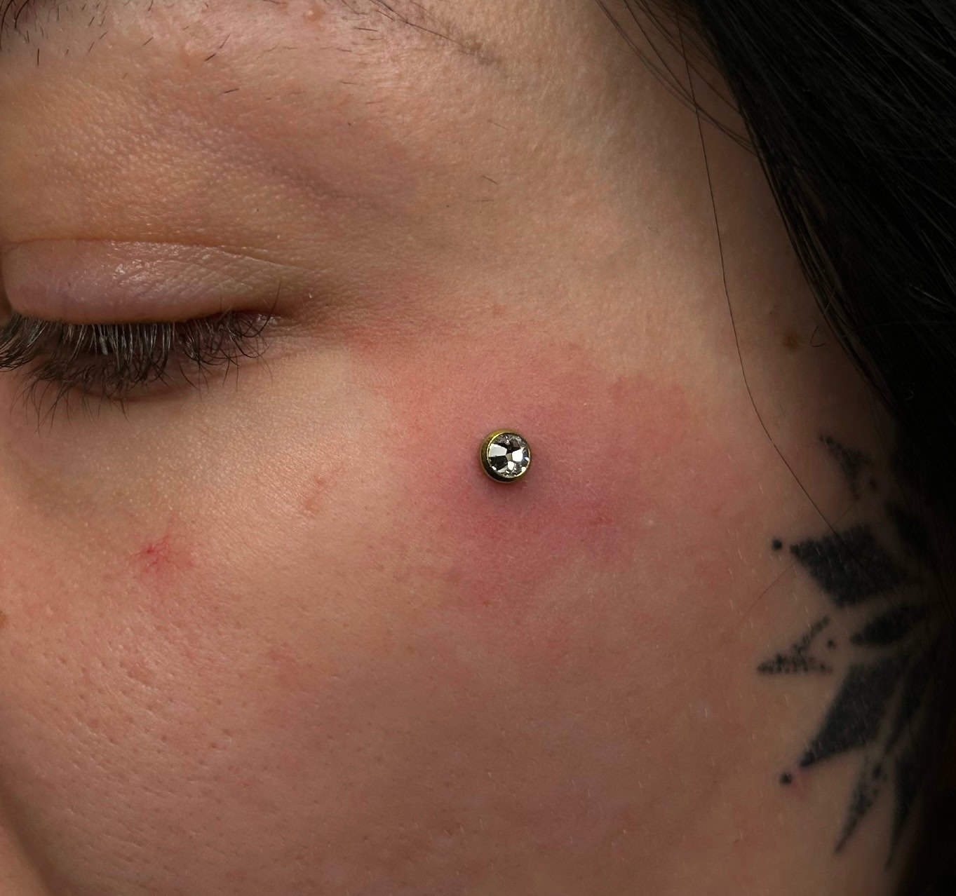 7sinstattoo.pl Zbliżenie na policzek osoby pokazujące IMG_5683, piercing skórny z okrągłym, srebrnym klejnotem. Skóra wokół piercingu jest lekko zaczerwieniona. Część czarnego geometrycznego tatuażu jest widoczna w pobliżu piercingu, prezentując kunszt studia tatuaży Poznań, a włosy osoby są ciemne. Oko osoby jest zamknięte. Najlepsze studio tatuażu Seven Sins Tattoo