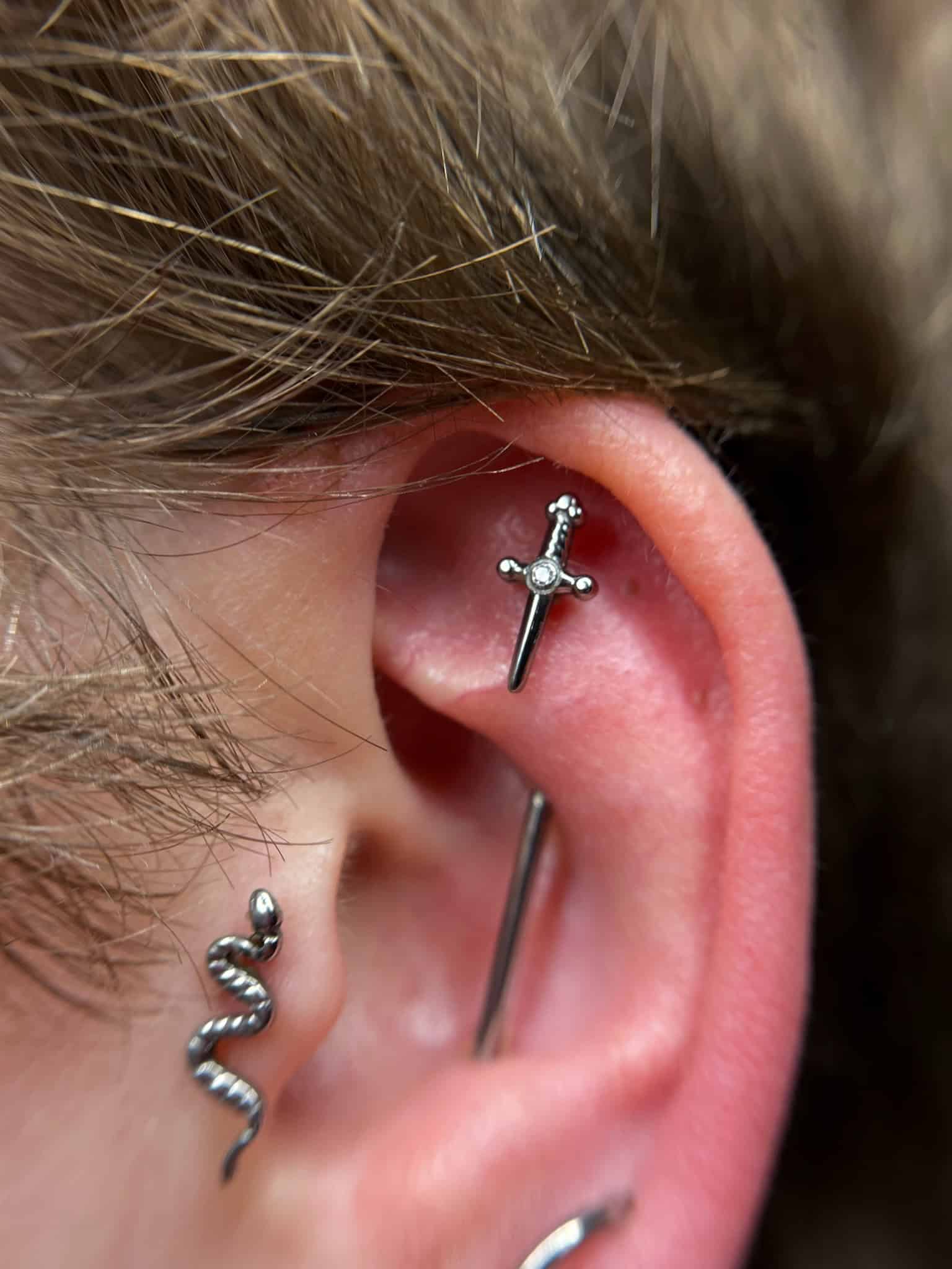 7sinstattoo.pl Zbliżenie ucha z trzema piercingami zaprezentowanymi w najlepszym studio tatuażu w Poznaniu. Piercingi obejmują mały kolczyk w kształcie miecza w chrząstce, srebrny kolczyk w kształcie sztangi nad nim i kolczyk w kształcie węża w płatku ucha. Jasne włosy są częściowo widoczne w tle. Odniesienie do obrazu: IMG_6075. Najlepsze studio tatuażu Seven Sins Tattoo