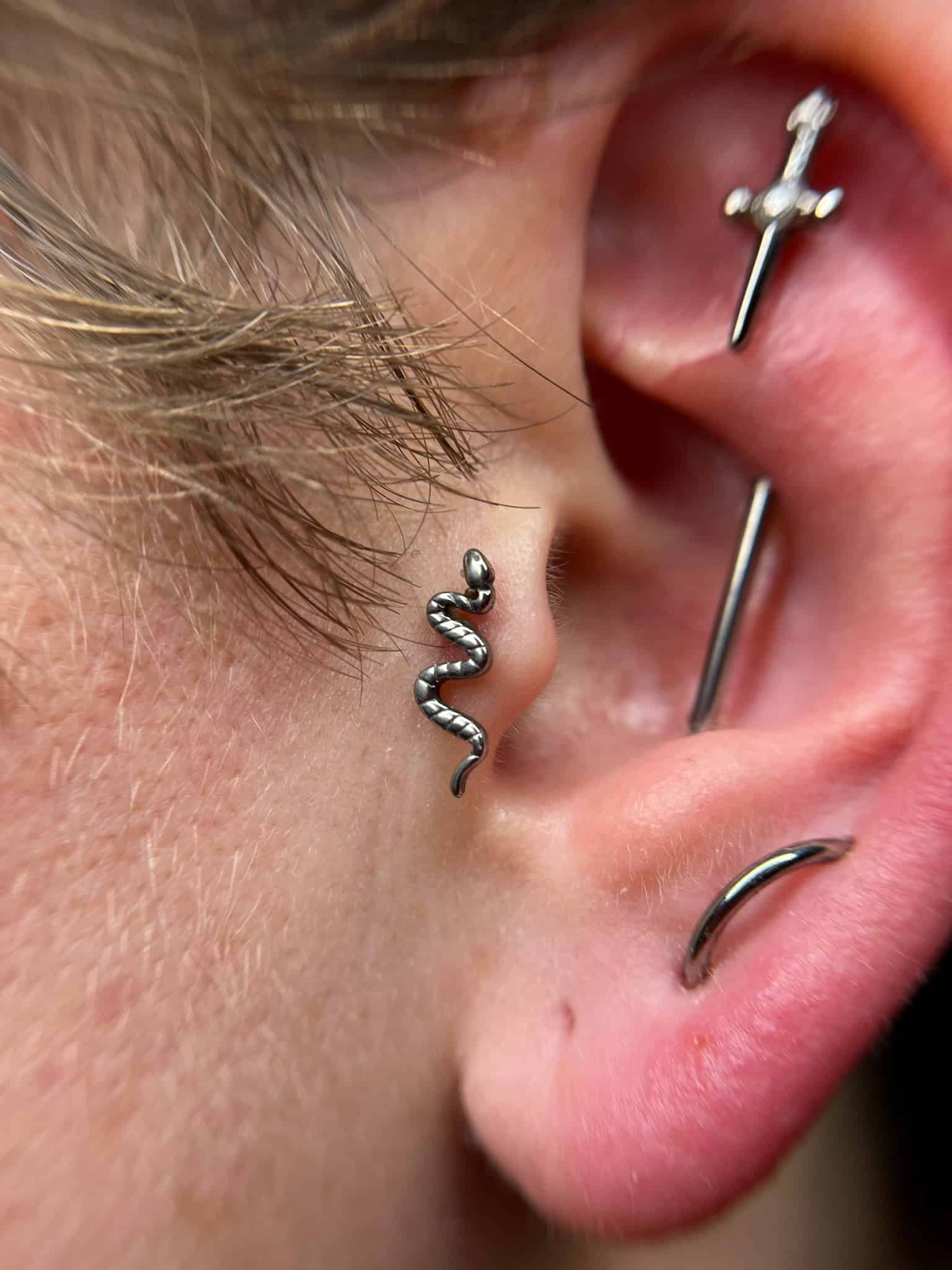 7sinstattoo.pl Zbliżenie ucha ozdobionego wieloma piercingami: małym kolczykiem w kształcie węża, kolczykiem w kształcie sztangi przypominającym miecz i kółkiem. Skóra wokół tych artystycznych elementów jest lekko czerwona. Odkryj więcej wyjątkowych stylów takich jak ten w Najlepszym Studio Tatuażu Poznań, znanym z wyjątkowej sztuki na ciele. (Nazwa produktu: IMG_6076) Najlepsze studio tatuażu Seven Sins Tattoo
