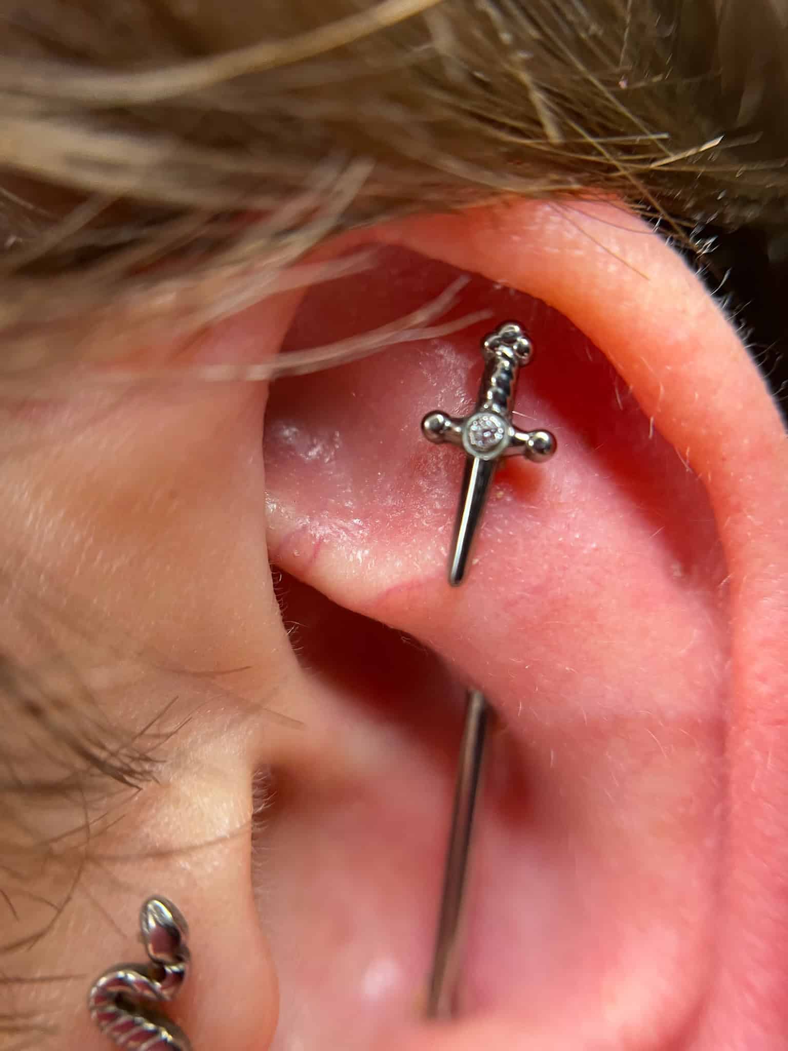 7sinstattoo.pl Zbliżenie na ucho z dwoma piercingami z najlepszego studia tatuażu Poznań (IMG_6092). Górny piercing ma srebrny kolczyk w kształcie małego miecza z klejnotem na rękojeści. Dolny piercing ma kolczyk w kształcie kółka ozdobiony zwiniętym wąż. Włosy częściowo widoczne po lewej stronie. Najlepsze studio tatuażu Seven Sins Tattoo