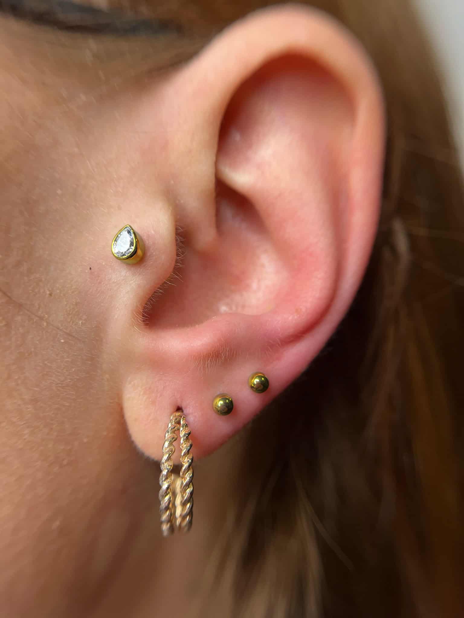 7sinstattoo.pl Zbliżenie ucha ozdobionego wieloma piercingami ze studia tatuaży Poznań. Ucho ma mały, przezroczysty kolczyk z kamieniem szlachetnym u góry, dwa małe złote kolczyki kulkowe na płatku ucha i złoty kolczyk-kółko ze skręconym wzorem poniżej płatka ucha, uchwycony w produkcie IMG_6132. Najlepsze studio tatuażu Seven Sins Tattoo