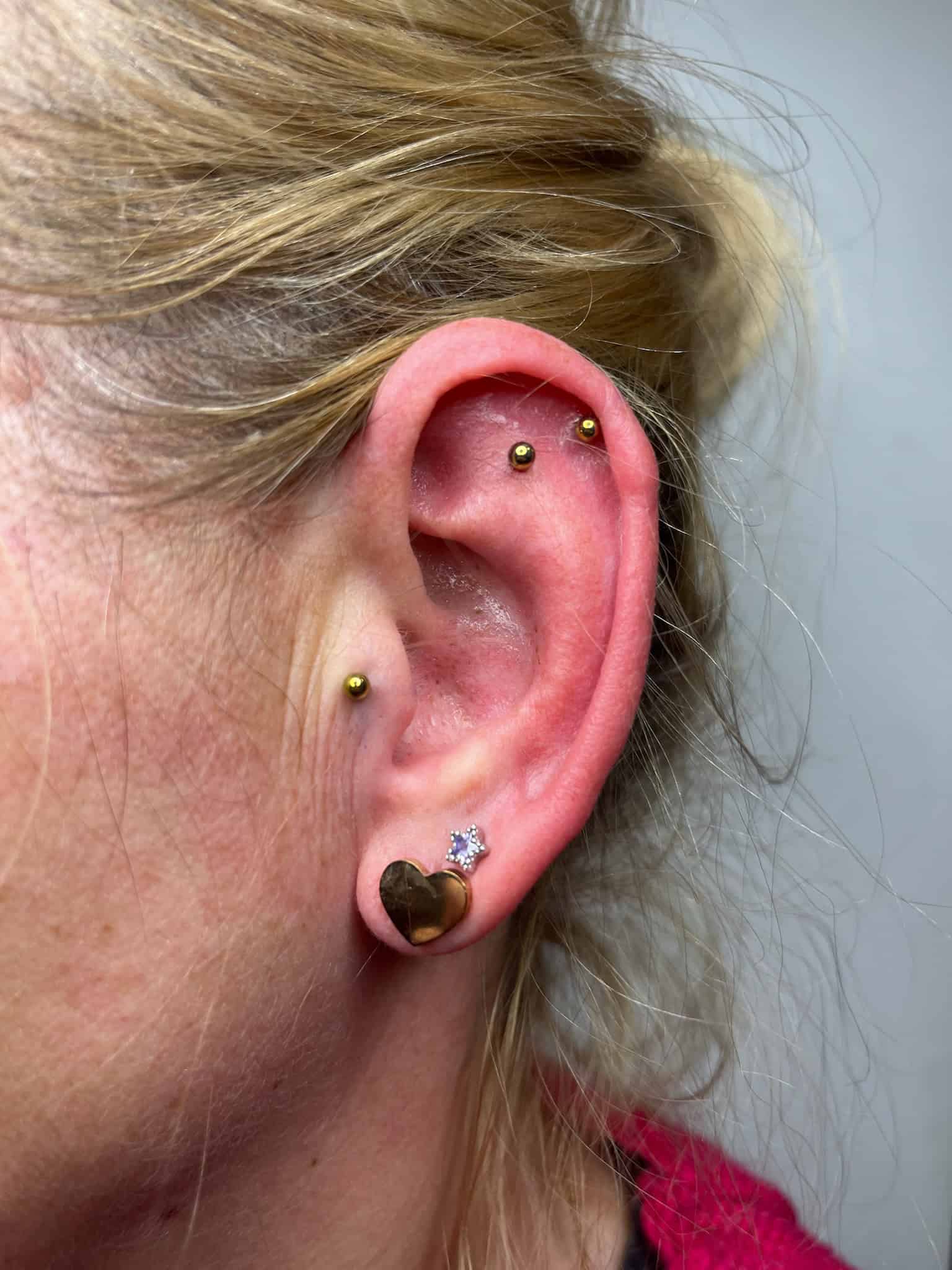 7sinstattoo.pl Zbliżenie na zewnętrzne ucho osoby z wieloma piercingami, zrobione w studio tatuażu Poznań. Ucho prezentuje obraz IMG_6205, ze złotym kolczykiem na płatku, jednym złotym piercingiem z klejnotem w kształcie gwiazdy i dwoma złotymi okrągłymi sztangami na górnej części. Osoba ma blond włosy związane z tyłu i nosi czerwony strój. Najlepsze studio tatuażu Seven Sins Tattoo