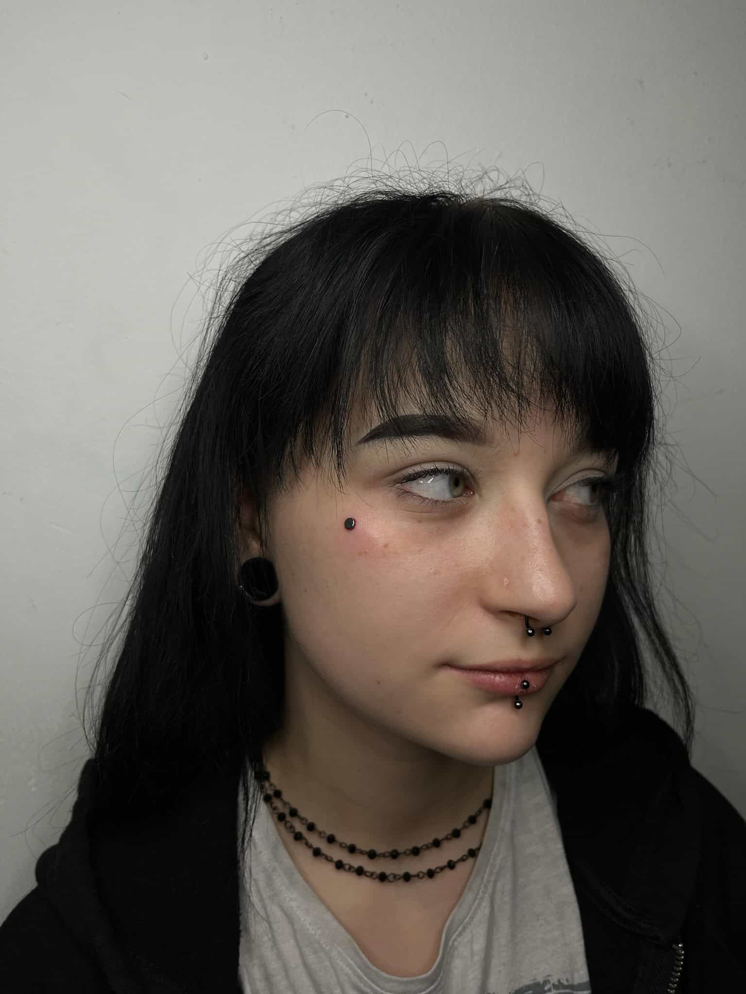 7sinstattoo.pl IMG_6365 przedstawia osobę z długimi czarnymi włosami, ciemną grzywką i wieloma piercingami twarzy, w tym piercingiem brwi, nosa i warg. Patrzy w bok, mając na sobie czarny kaptur i czarne naszyjniki z koralików. Osoba ta zachowuje neutralny wyraz twarzy przed jednolitym tłem w studio tatuażu Poznań. Najlepsze studio tatuażu Seven Sins Tattoo