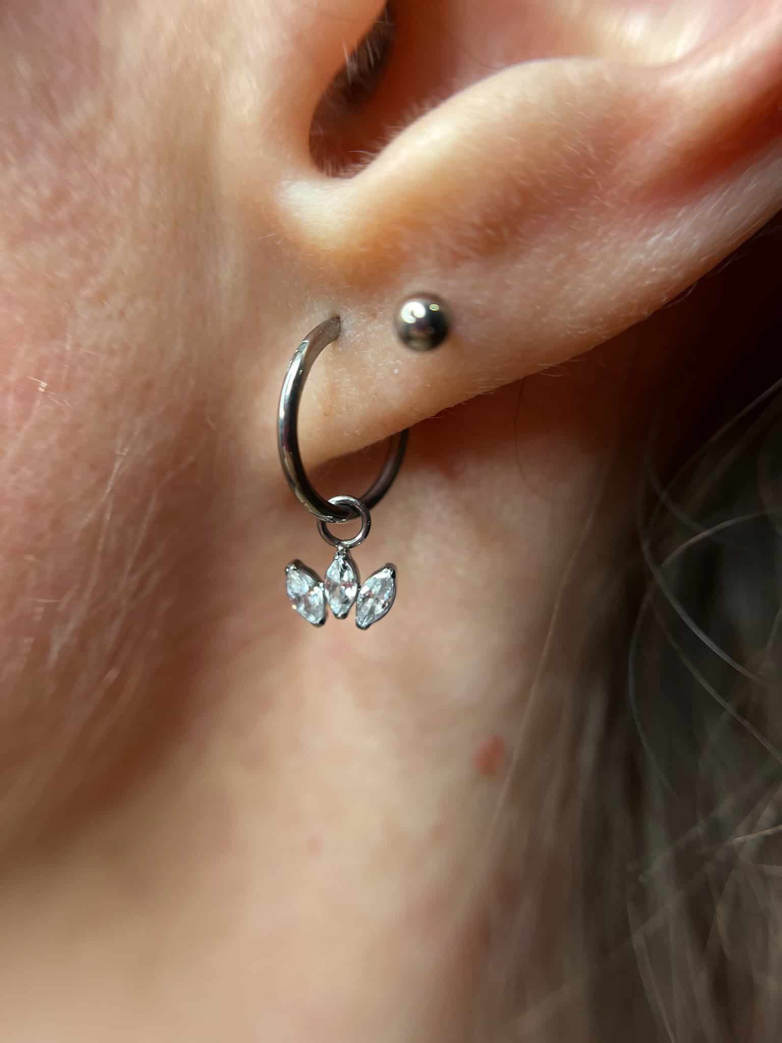 7sinstattoo.pl Zbliżenie ucha ozdobionego obrazem IMG_6660, z małym srebrnym kolczykiem w górnej części i oszałamiającym srebrnym kolczykiem w kształcie koła z dwoma wiszącymi kryształowymi motylami w dolnym płacie. Jasny odcień skóry uzupełnia kilka pasm włosów w tle, tworząc artystyczny efekt przypominający studio tatuaży Poznań. Najlepsze studio tatuażu Seven Sins Tattoo