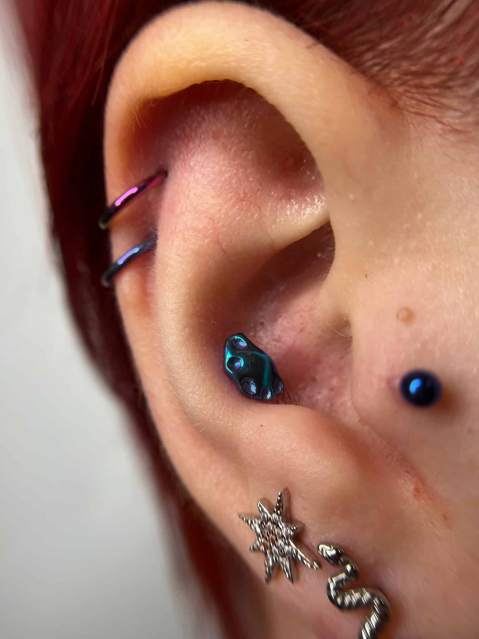 7sinstattoo.pl Zbliżenie ucha osoby ozdobionego wieloma kolczykami. Ucho ma pięć odrębnych elementów biżuterii, w tym dwa kółka u góry, mały niebieski kolczyk w środku ucha, kolczyk w kształcie gwiazdy na dolnej części i kolczyk w kształcie węża na dole — idealny do pokazania się po wizycie w tatuaż Poznań. Nazwa produktu: IMG_6668. Najlepsze studio tatuażu Seven Sins Tattoo