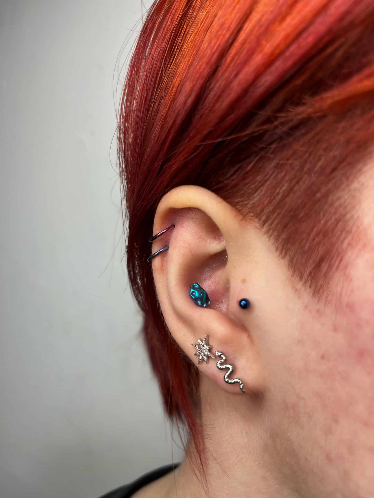 7sinstattoo.pl Zbliżenie ucha osoby z wieloma piercingami i jaskrawoczerwonymi włosami. Ucho ma różnorodną biżuterię, w tym niebieski kolczyk w tragusie, mały niebieski kółko w helisie, srebrny kolczyk w kształcie węża i kolczyk w kształcie gwiazdy na płatku ucha. Odkryj więcej wyjątkowych stylów w Studio Tatuażu Poznań z IMG_6681. Najlepsze studio tatuażu Seven Sins Tattoo
