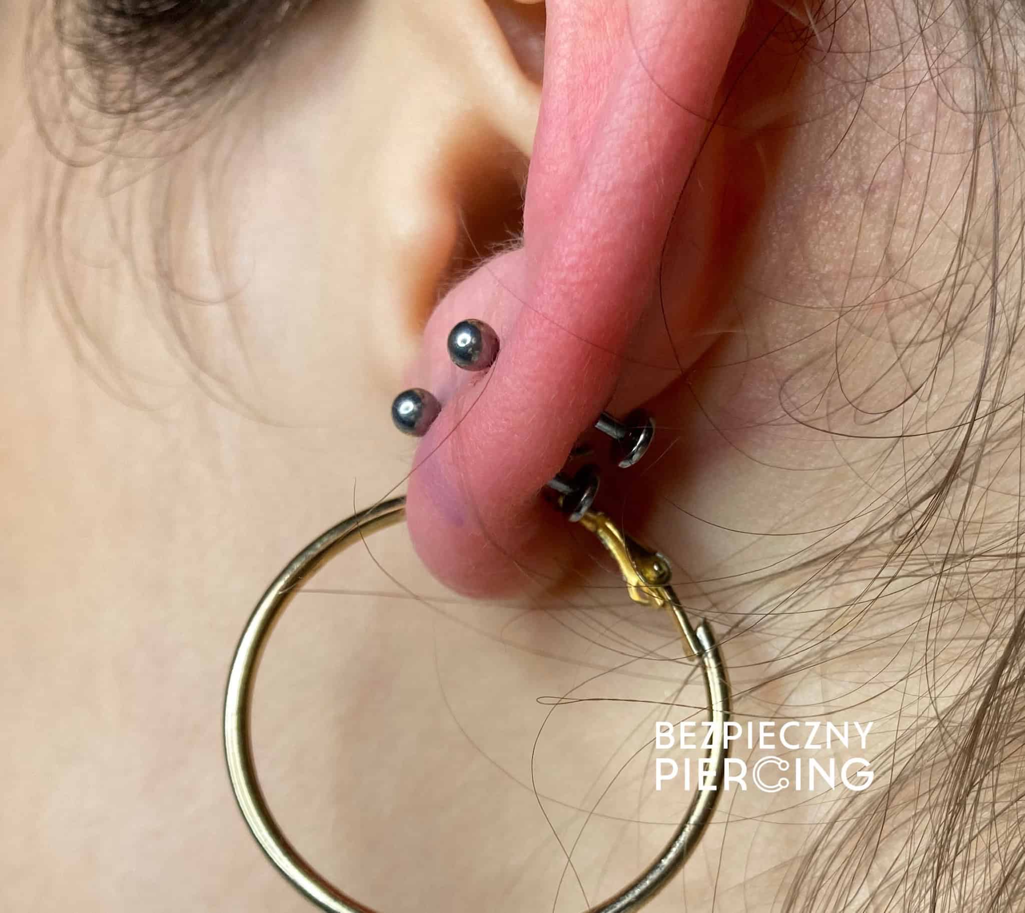 7sinstattoo.pl Zbliżenie ucha osoby ozdobionego dużym piercingiem i dwoma mniejszymi piercingami nad nim, wszystkie prezentujące stylową biżuterię. Wyraźny kolczyk-kółko jest osadzony w dużym piercingu. Tekst na zdjęciu, „Bezpieczny Piercing”, podkreśla studio tatuaży Poznań jako najlepszy wybór w zakresie modnej sztuki ciała. Produkt: IMG_6695 Najlepsze studio tatuażu Seven Sins Tattoo